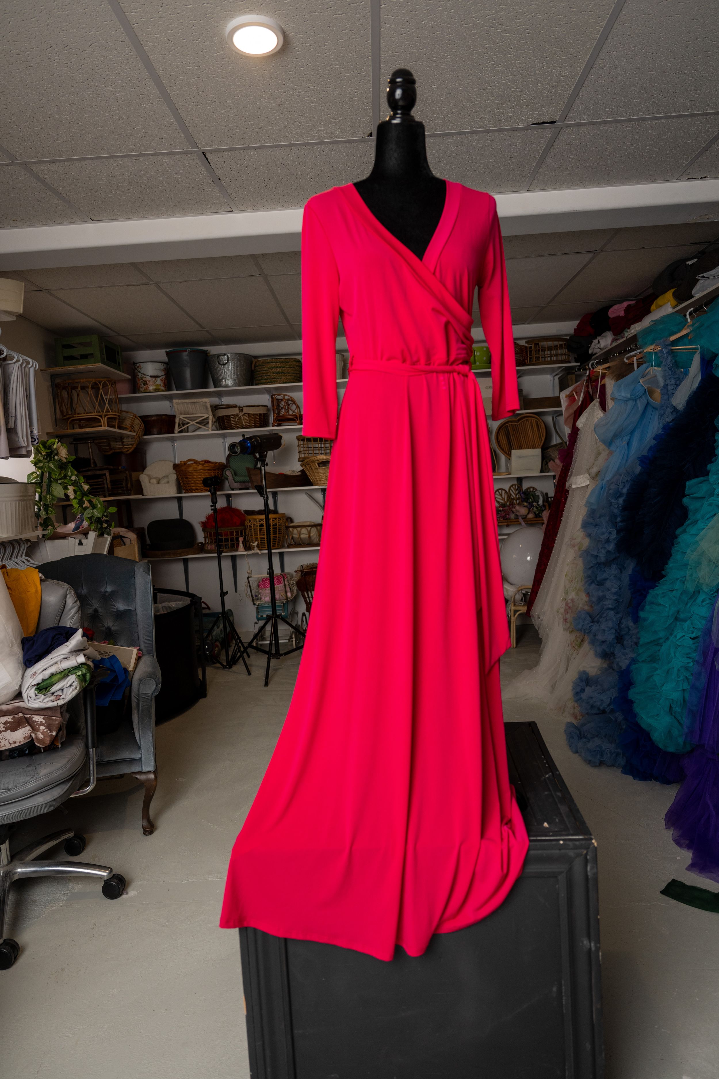 A bright red V-neck dress displayed on a black mannequin in a storage room with clothing racks and shelves.