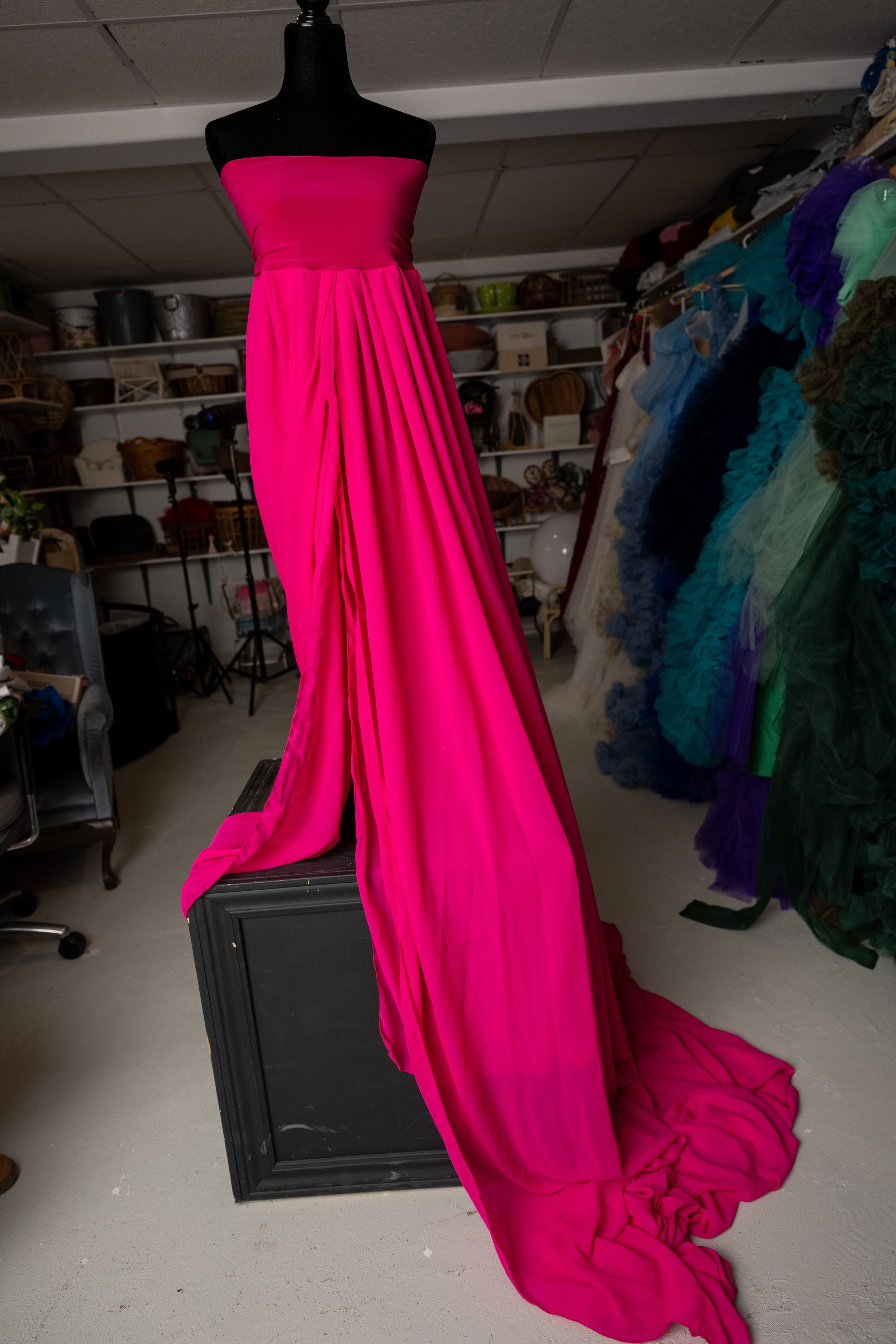 A vibrant hot pink evening gown with flowing fabric displayed on a black mannequin in a clothing studio setting.