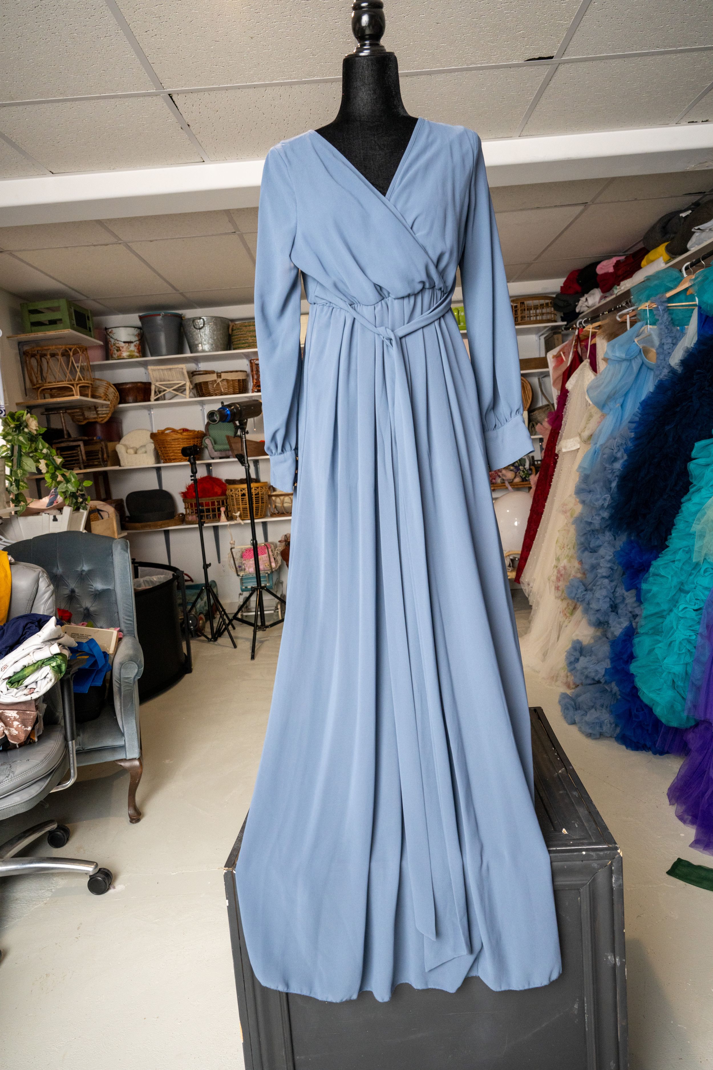 A light blue floor-length wrap dress with long sleeves displayed on a black mannequin in a clothing storage room.