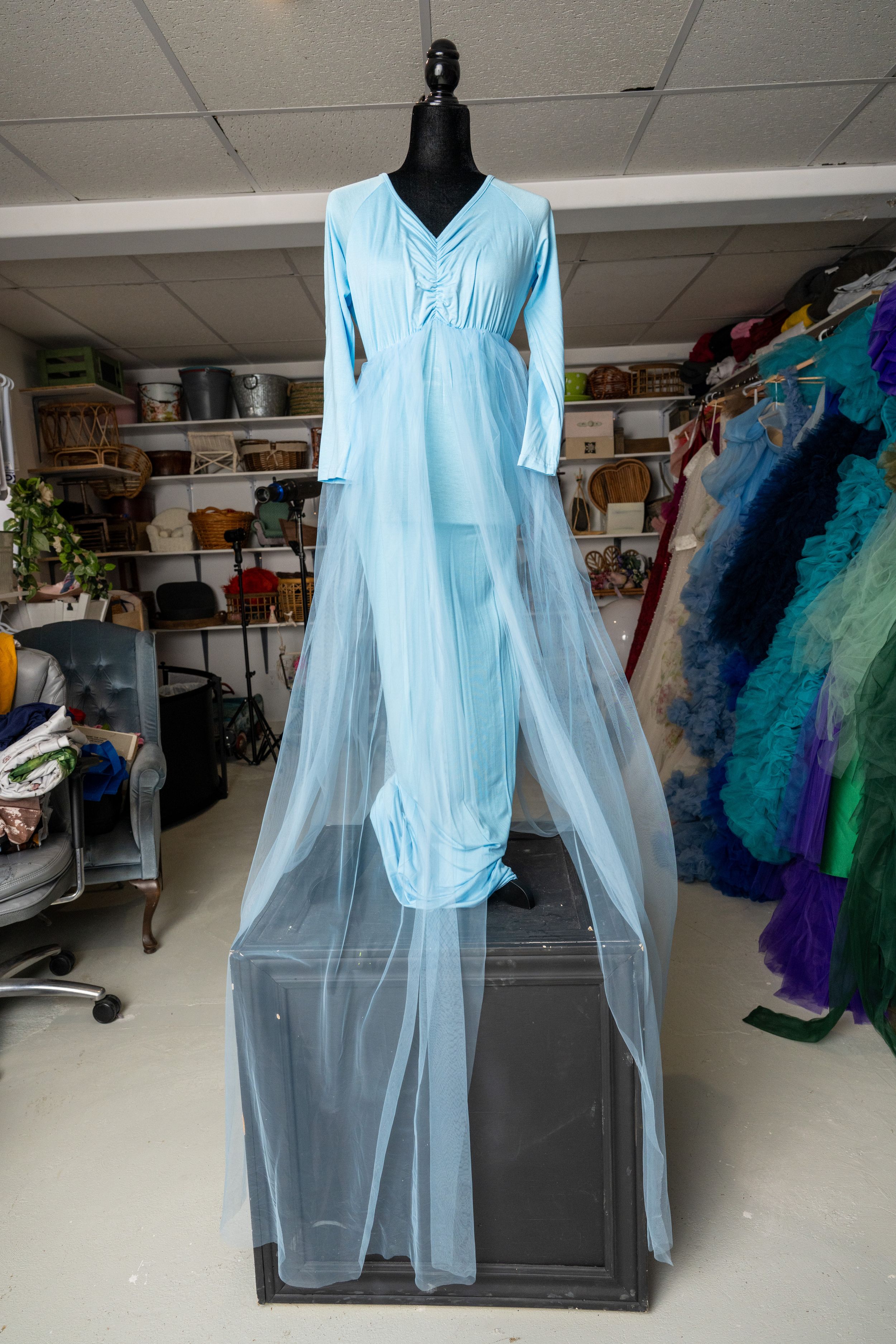 A light blue dress with sheer sleeves displayed on a mannequin in a storage room with colorful costumes in the background.
