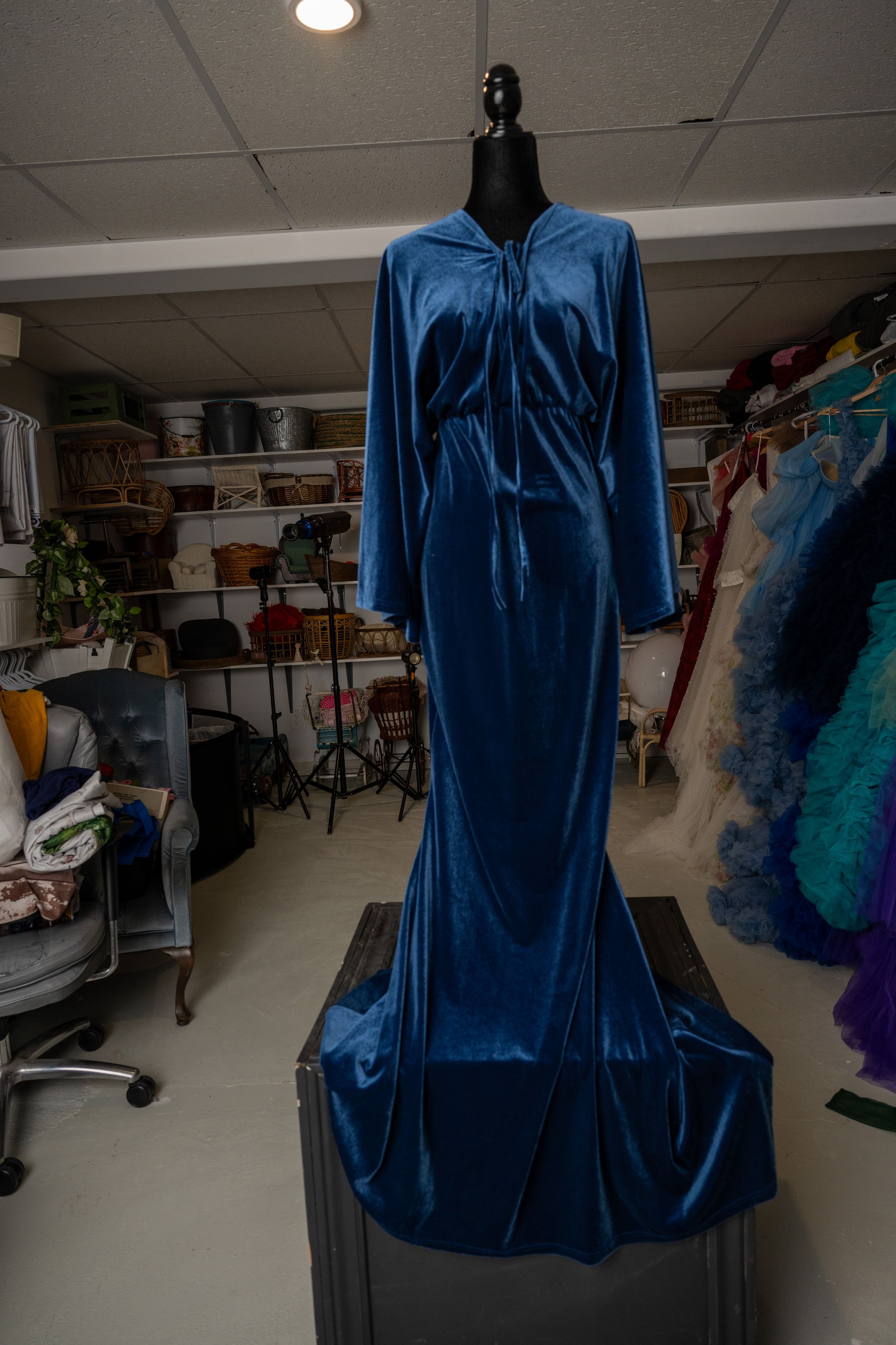 Blue velvet blouse displayed on mannequin in clothing workshop with storage and equipment visible in background.