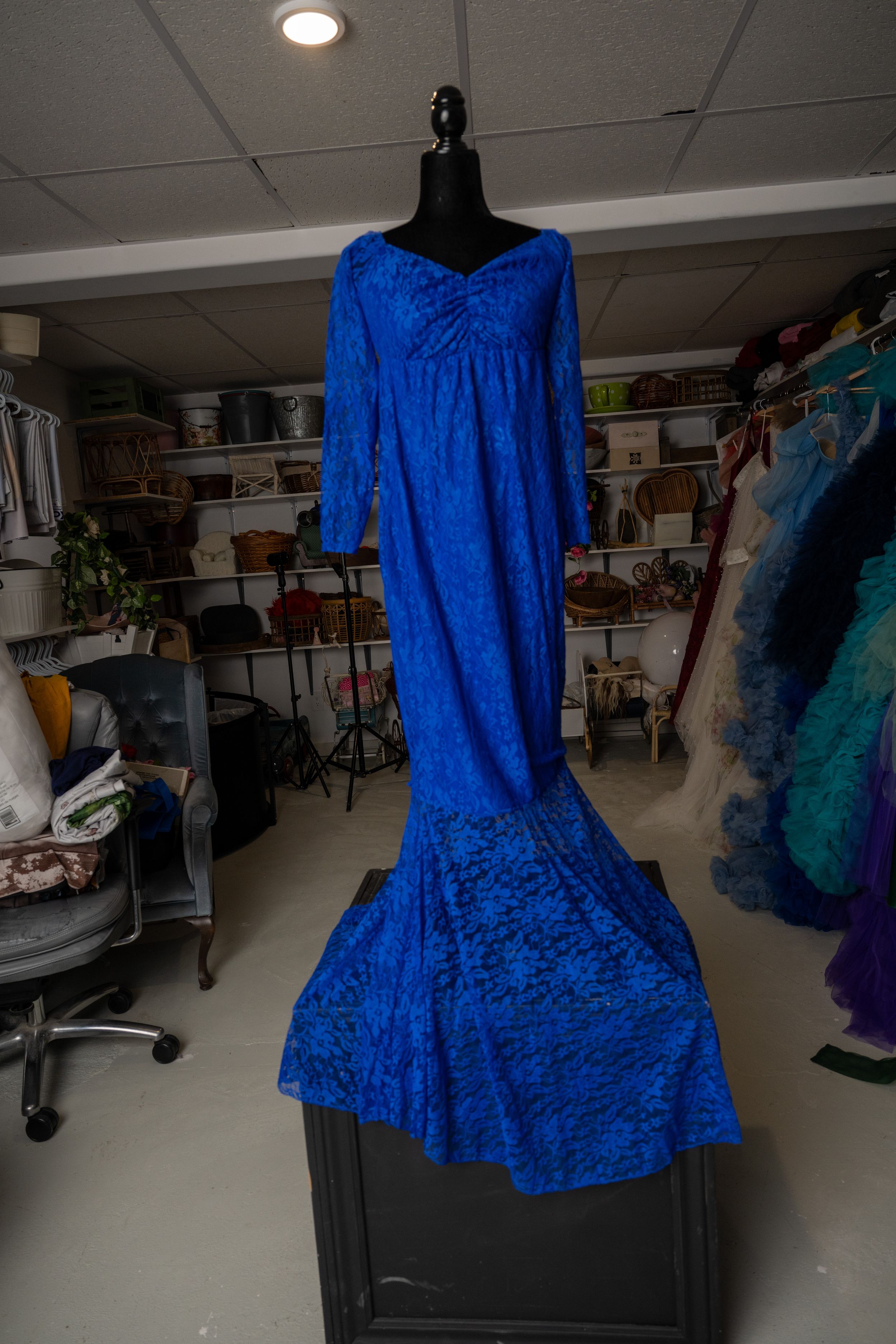 A vibrant royal blue lace dress with long sleeves and mermaid silhouette displayed on a black mannequin in a storage room.