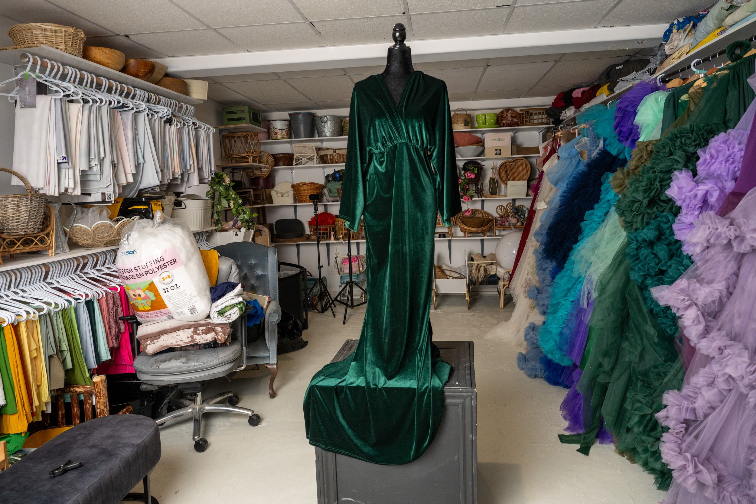A green graduation gown hangs in a storage room filled with costumes, clothing racks, and various theatrical supplies.