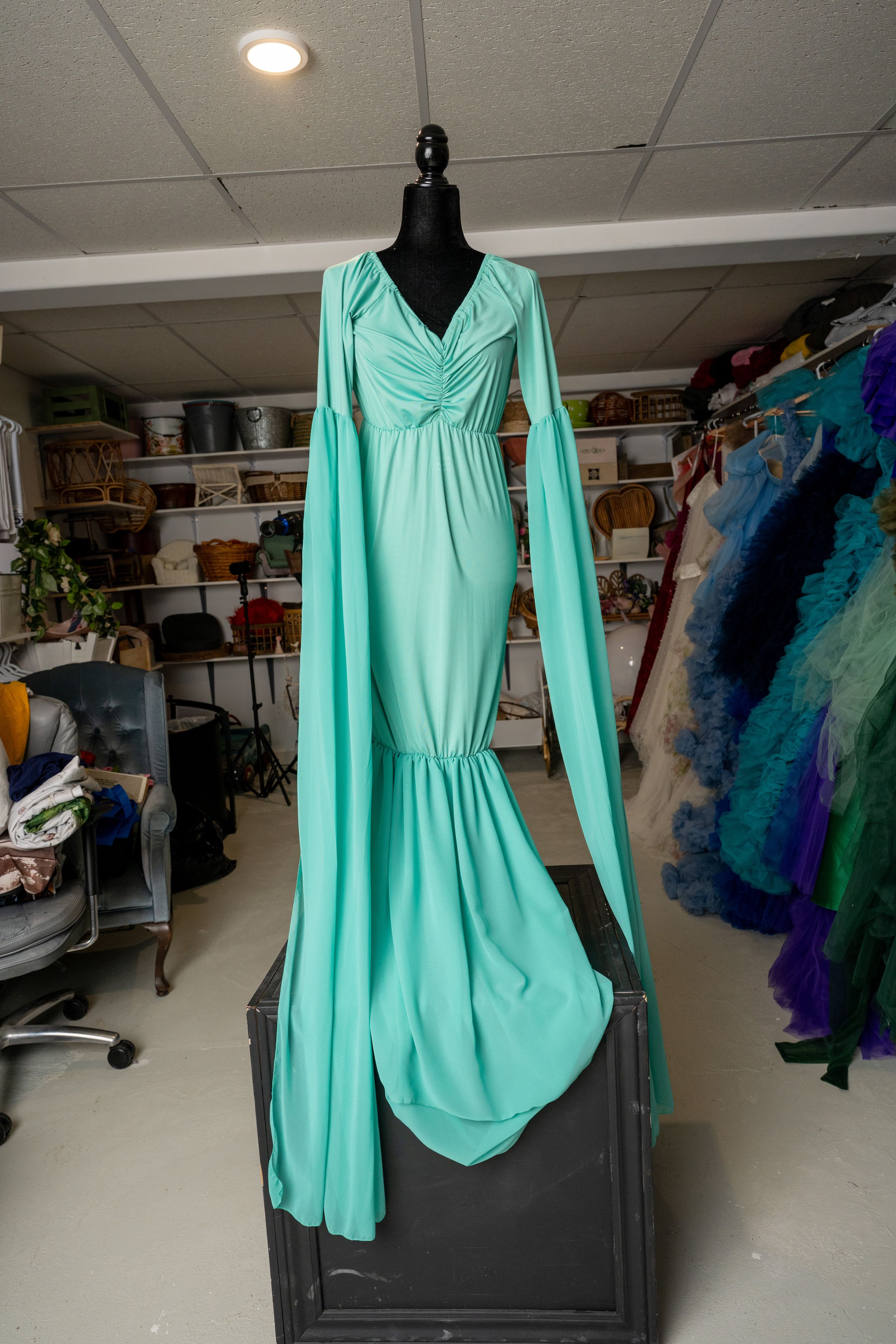 Mint green flowy dress with long sleeves displayed on a black mannequin in a storage room with colorful dresses visible.