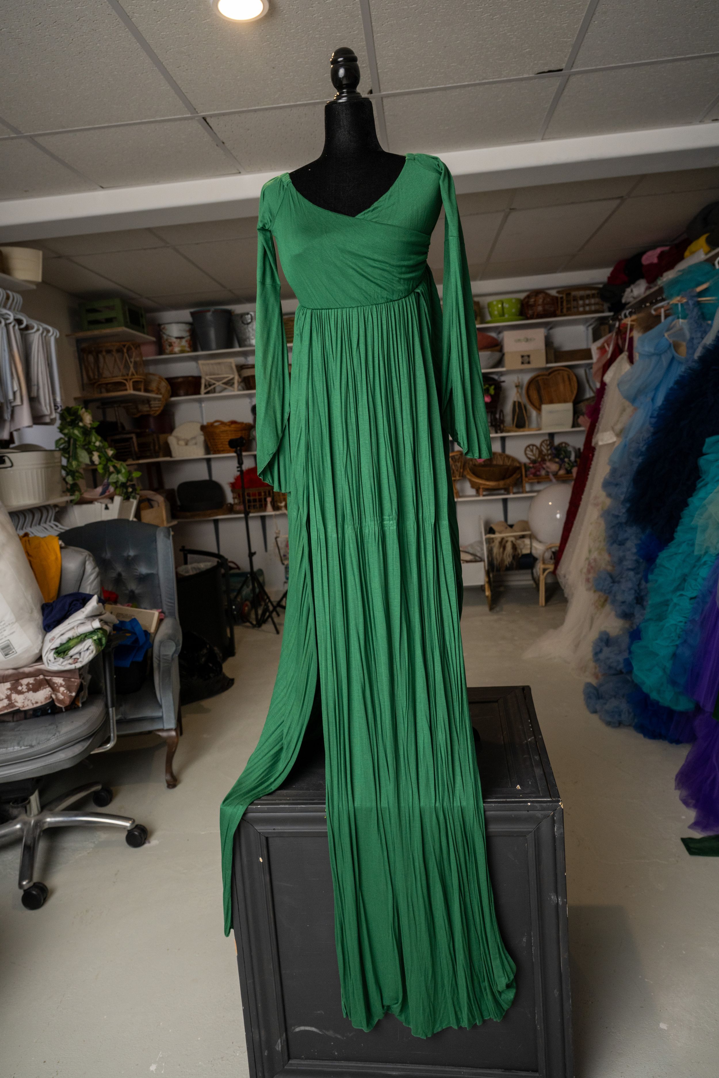 Kelly green pleated dress with long sleeves shown on a black dress form against a white wall with storage shelves.