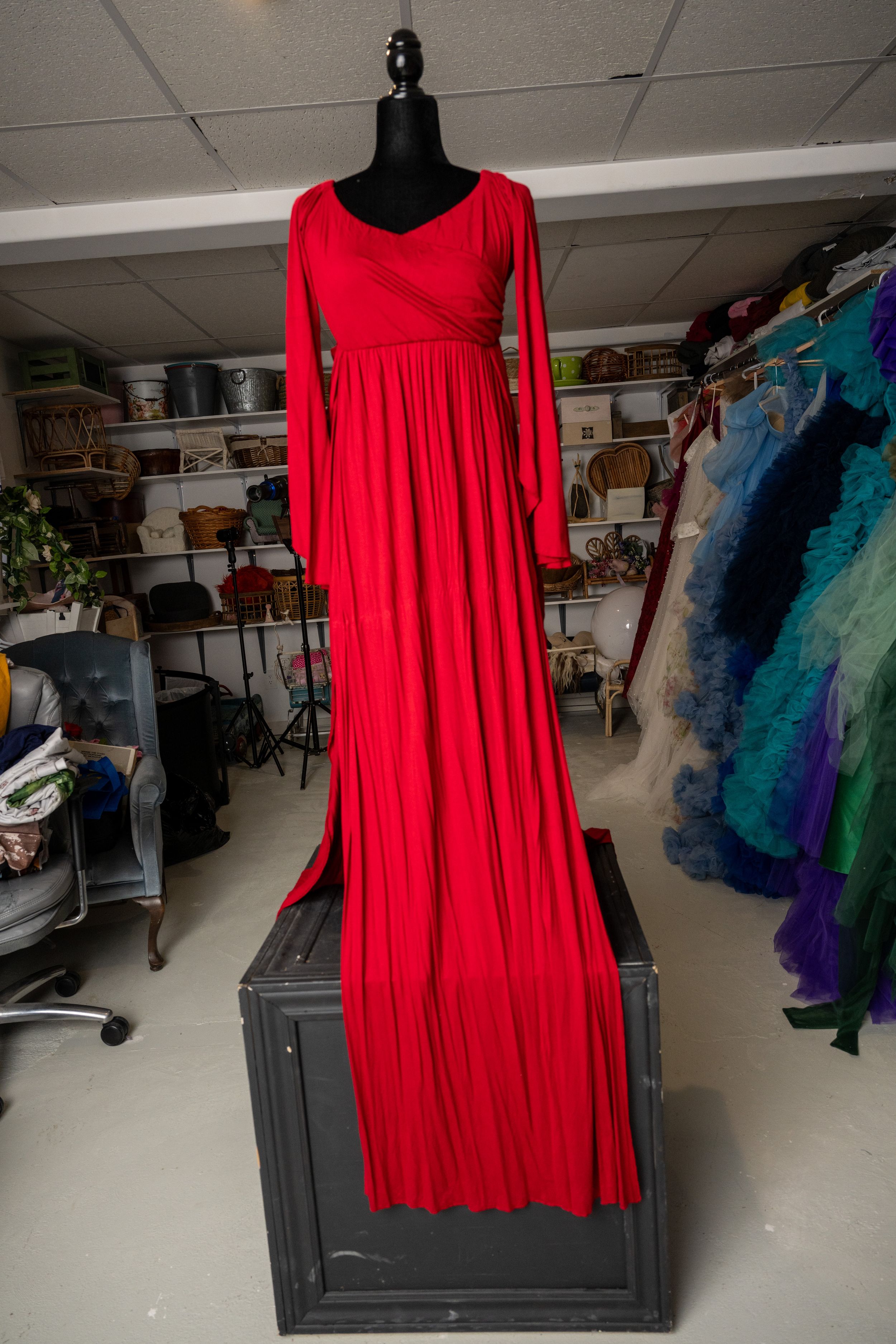 A bright red flowy dress displayed on a black mannequin in a storage room with shelving and colorful garments visible.