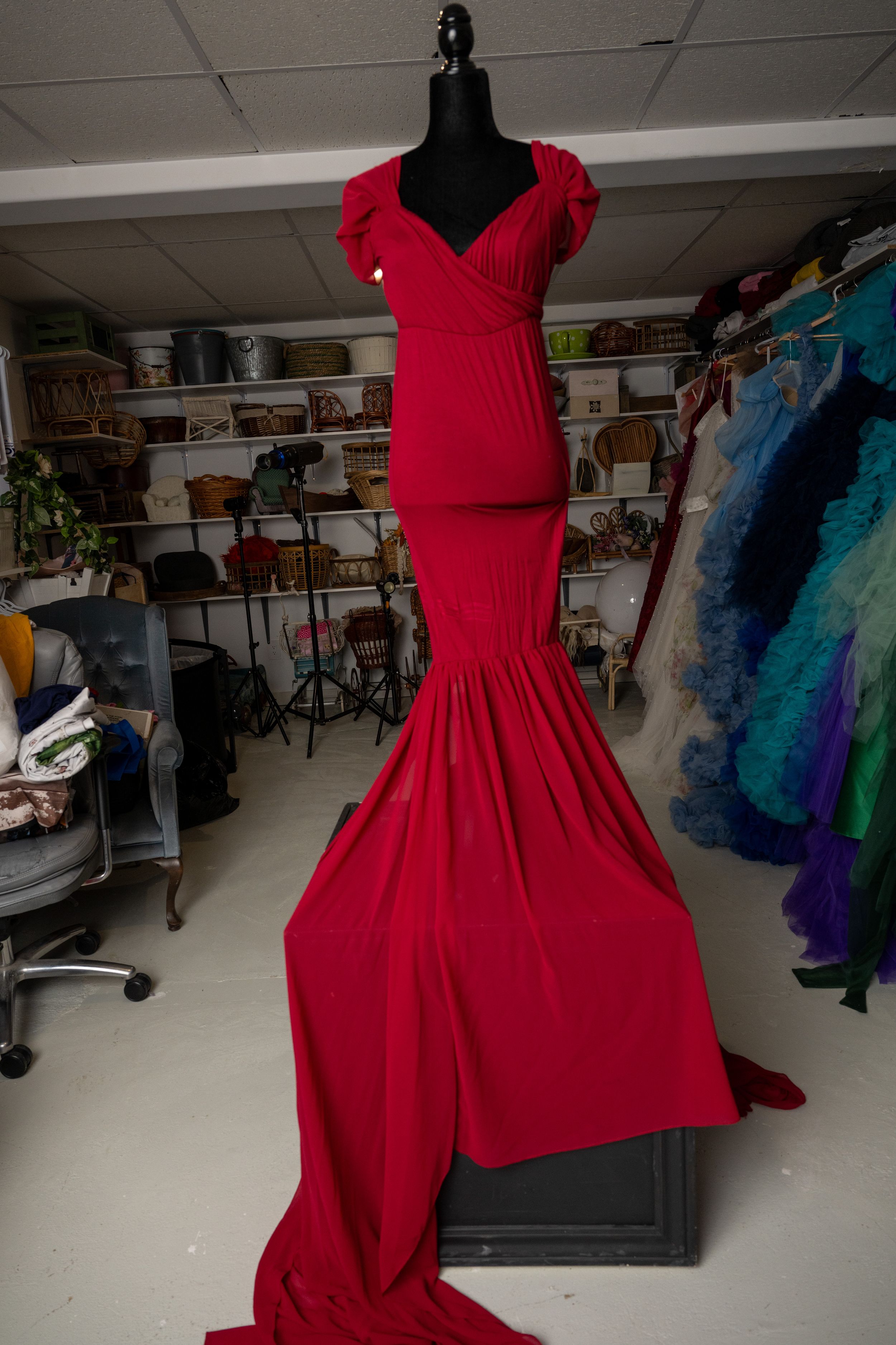 A red formal dress displayed on a black mannequin in a studio or workshop setting with other dresses visible in background.