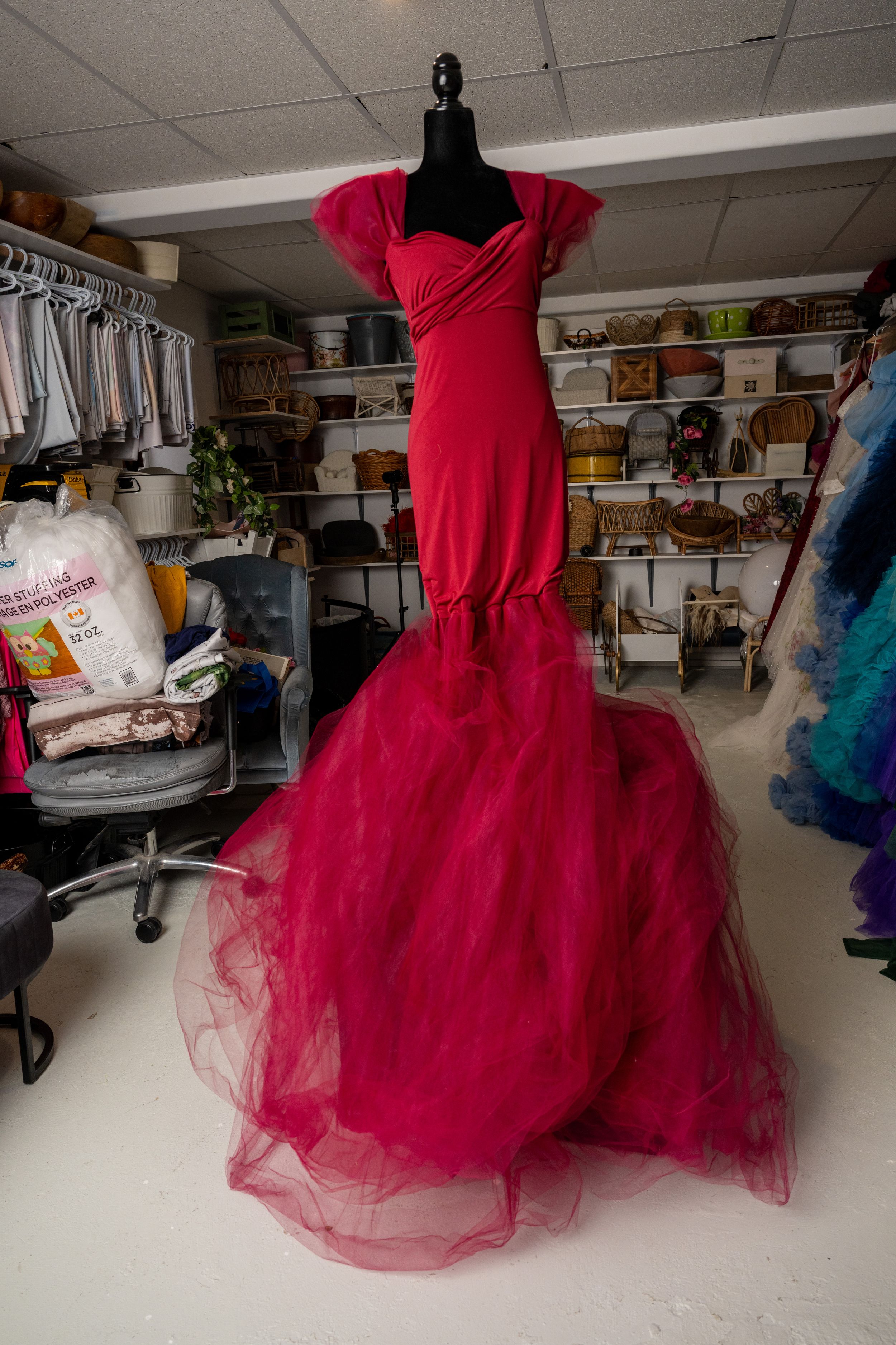A bright red formal gown with a voluminous tulle skirt is displayed on a dress form in a studio workspace.