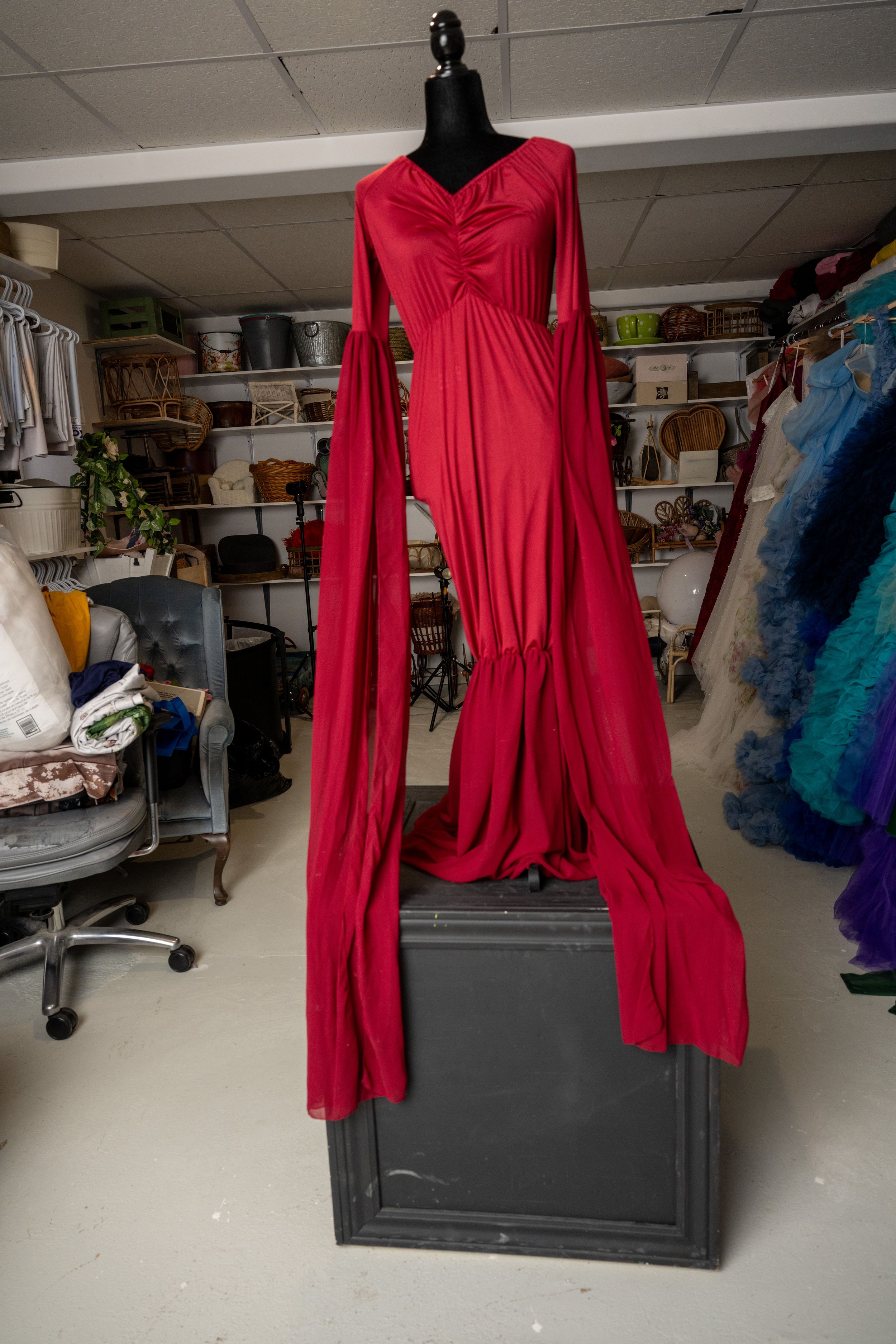 Red floor-length dress with long sleeves shown on a dress form in a workshop space with storage shelves.