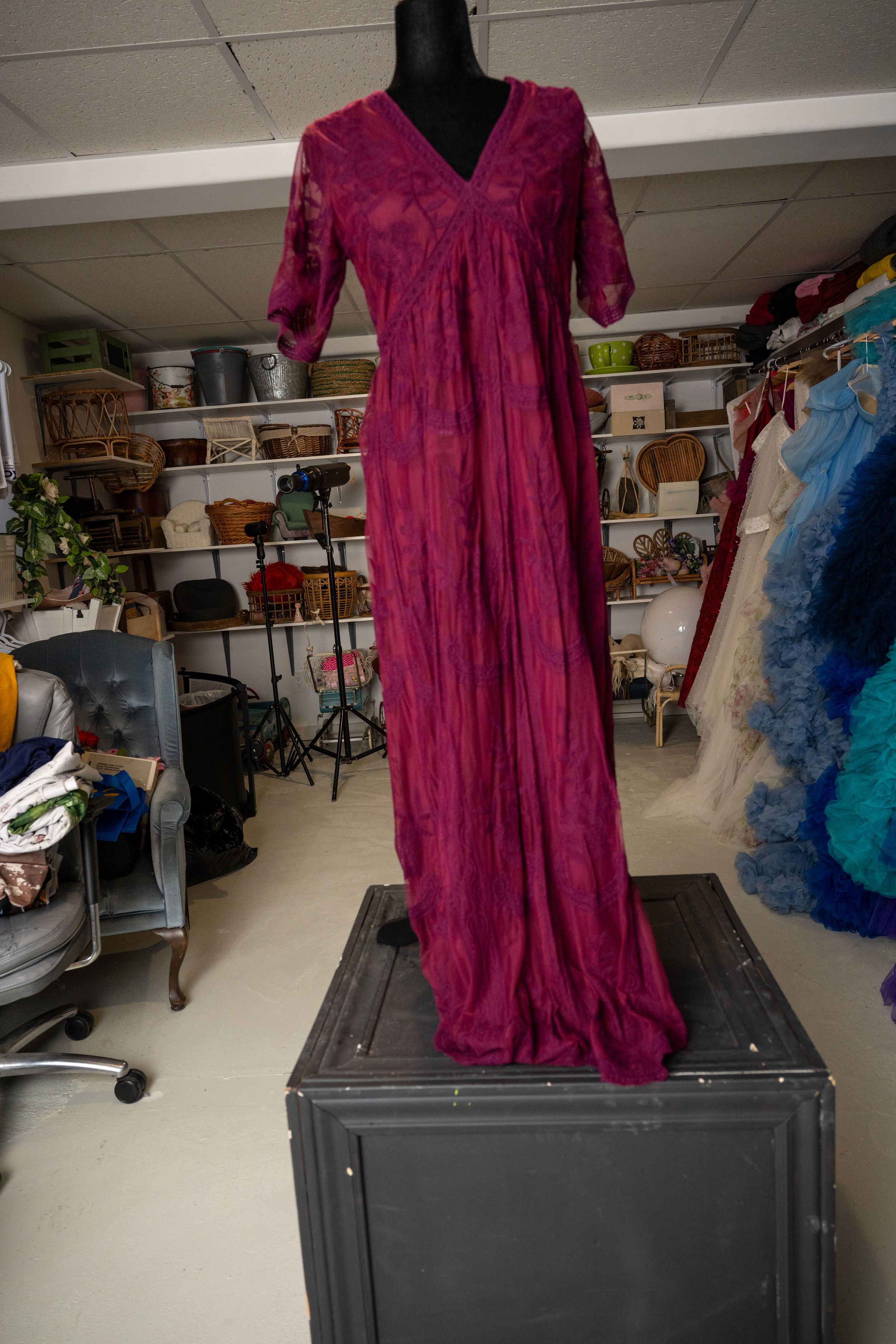 A burgundy velvet maxi dress with lace sleeves displayed on a mannequin in a cluttered storage room.