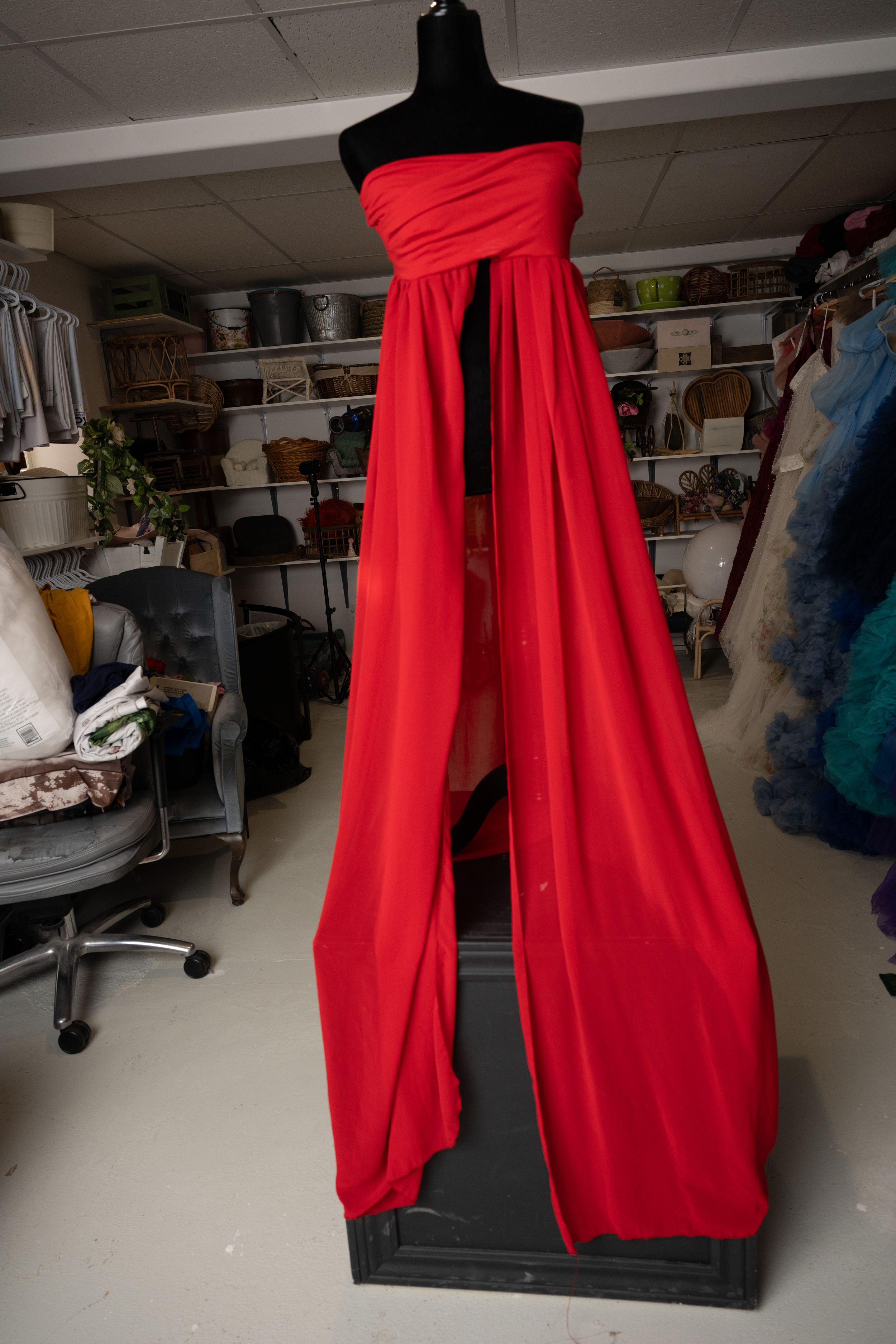 A red formal gown with draped fabric on a black mannequin stand displayed in a cluttered workshop or studio space.