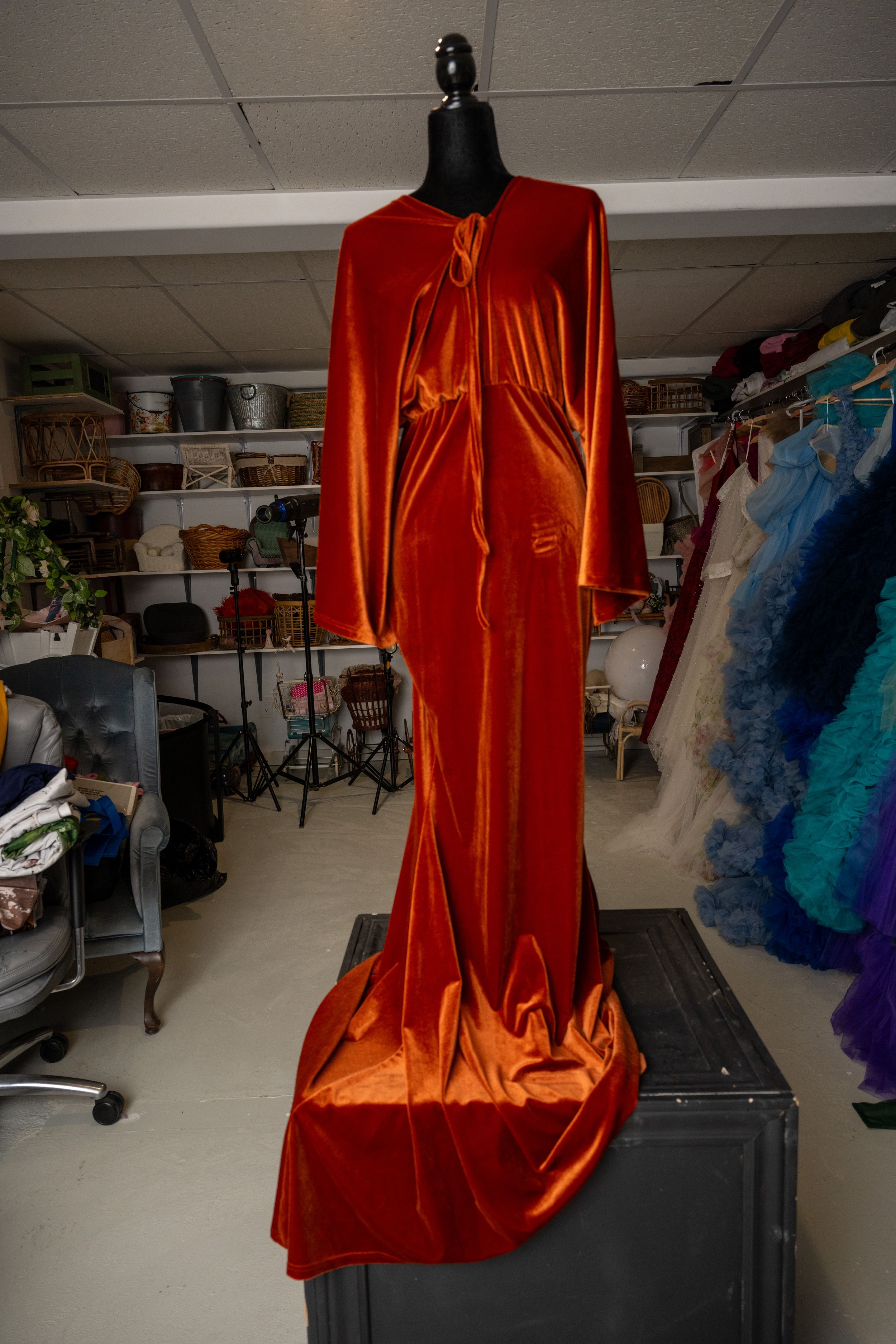 A long flowing burnt orange velvet gown with long sleeves displayed on a black mannequin in a costume storage room.