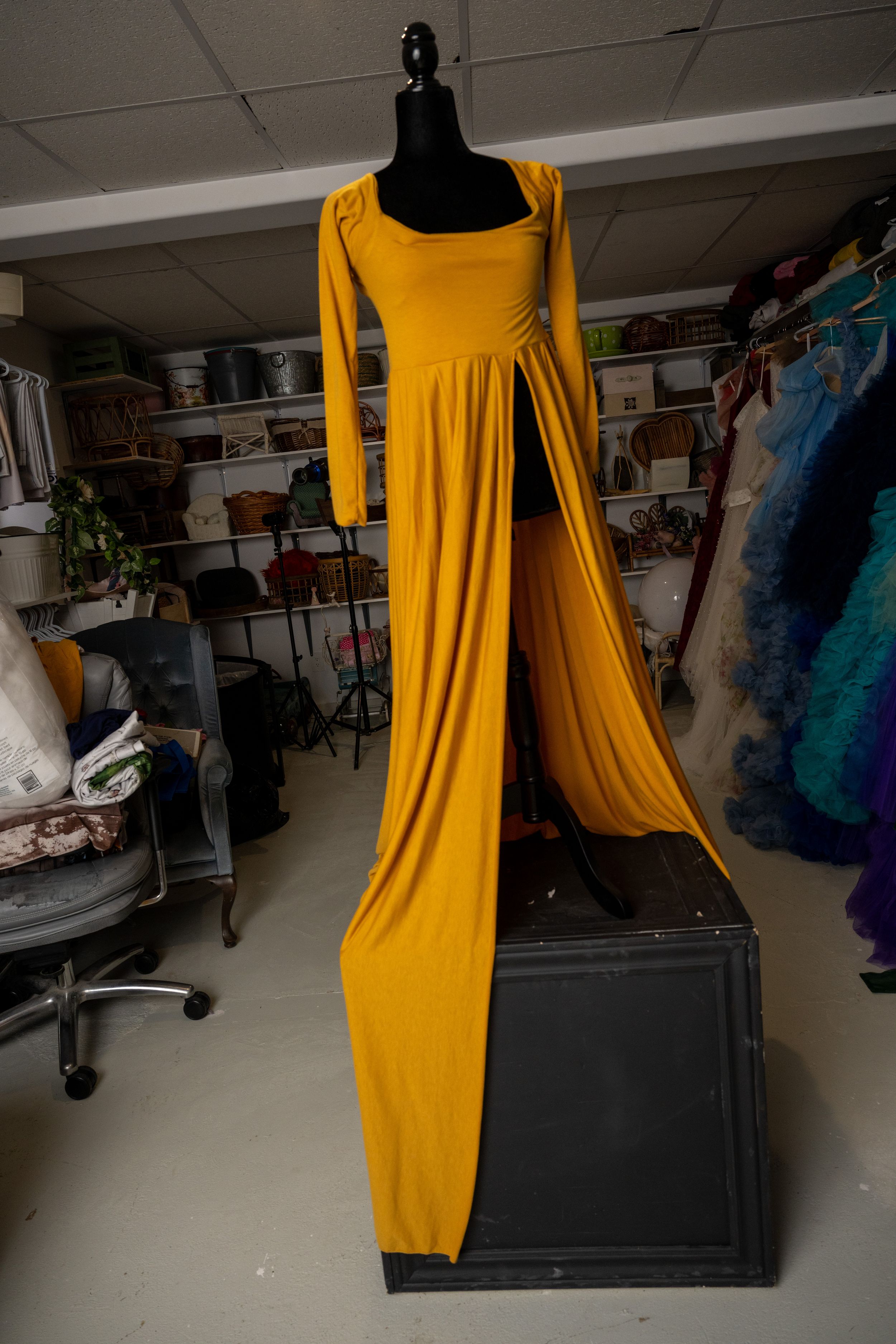A bright yellow dress with flowing cape sleeves is displayed on a black mannequin against a dark background.