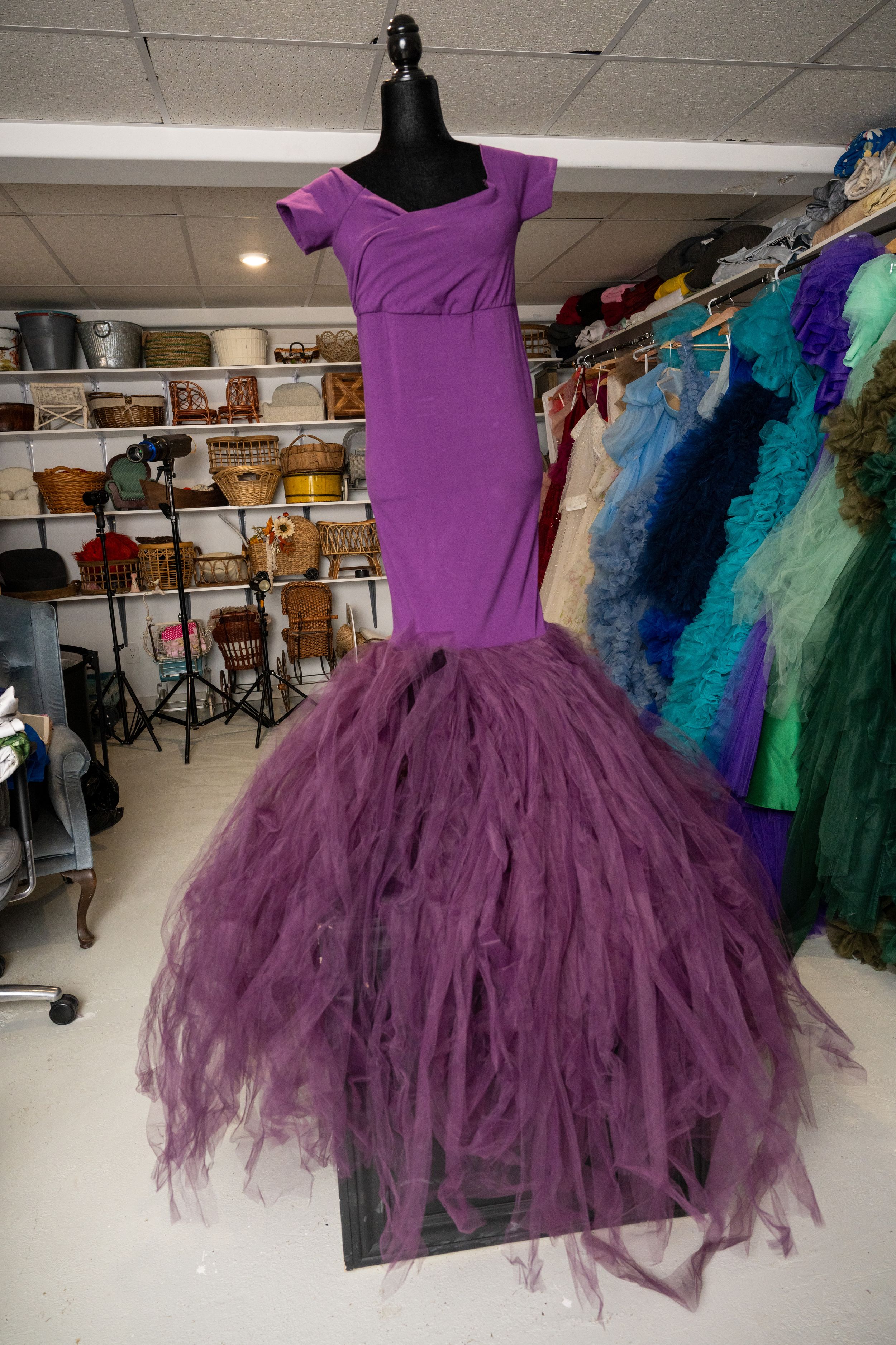 A purple tulle ballet dress with ruffled layers is displayed in a dance studio setting with other costumes visible.