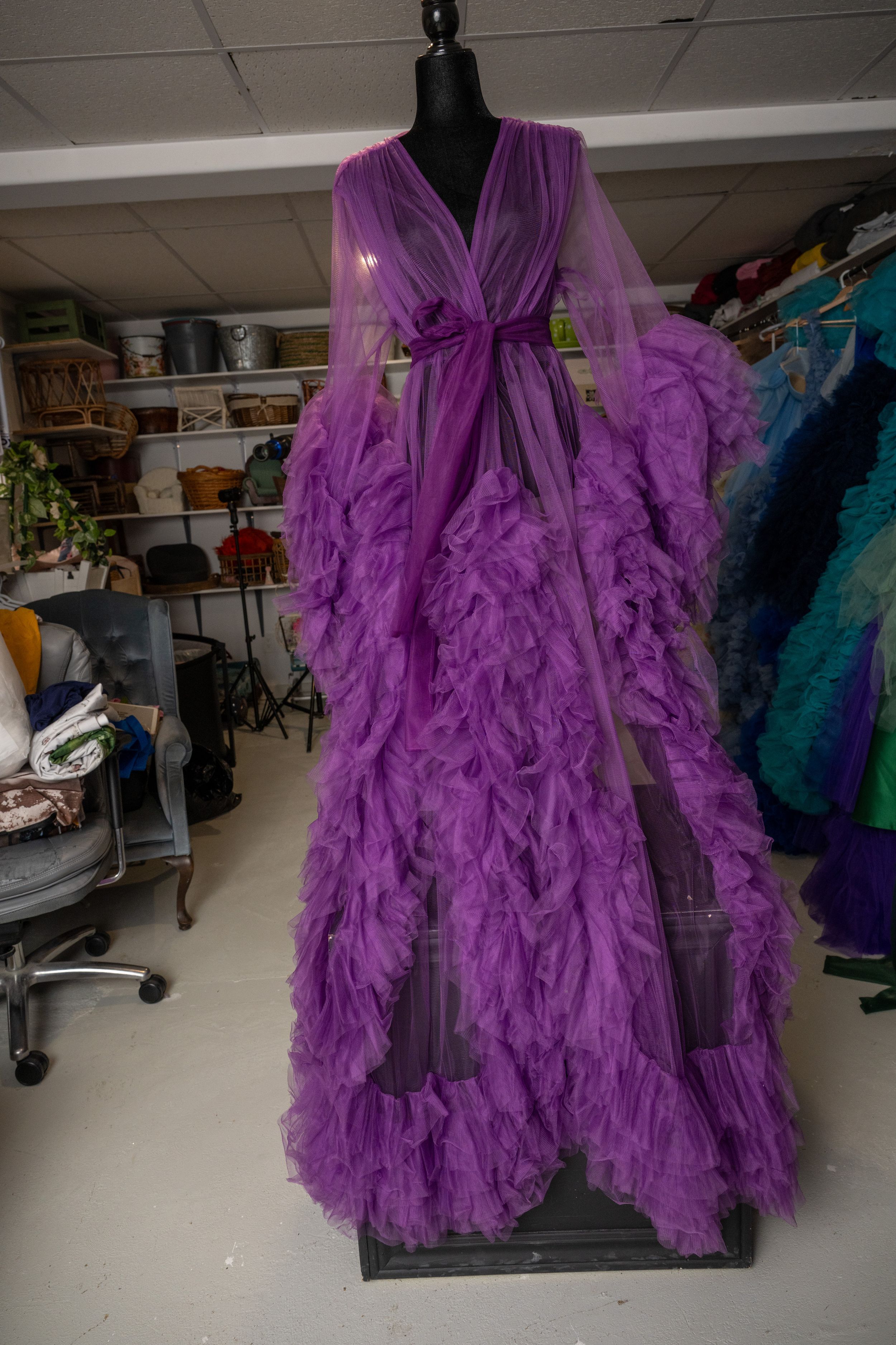 A flowing purple tulle dress with ruffled layers and bell sleeves shown on a dress form in a fashion workspace.