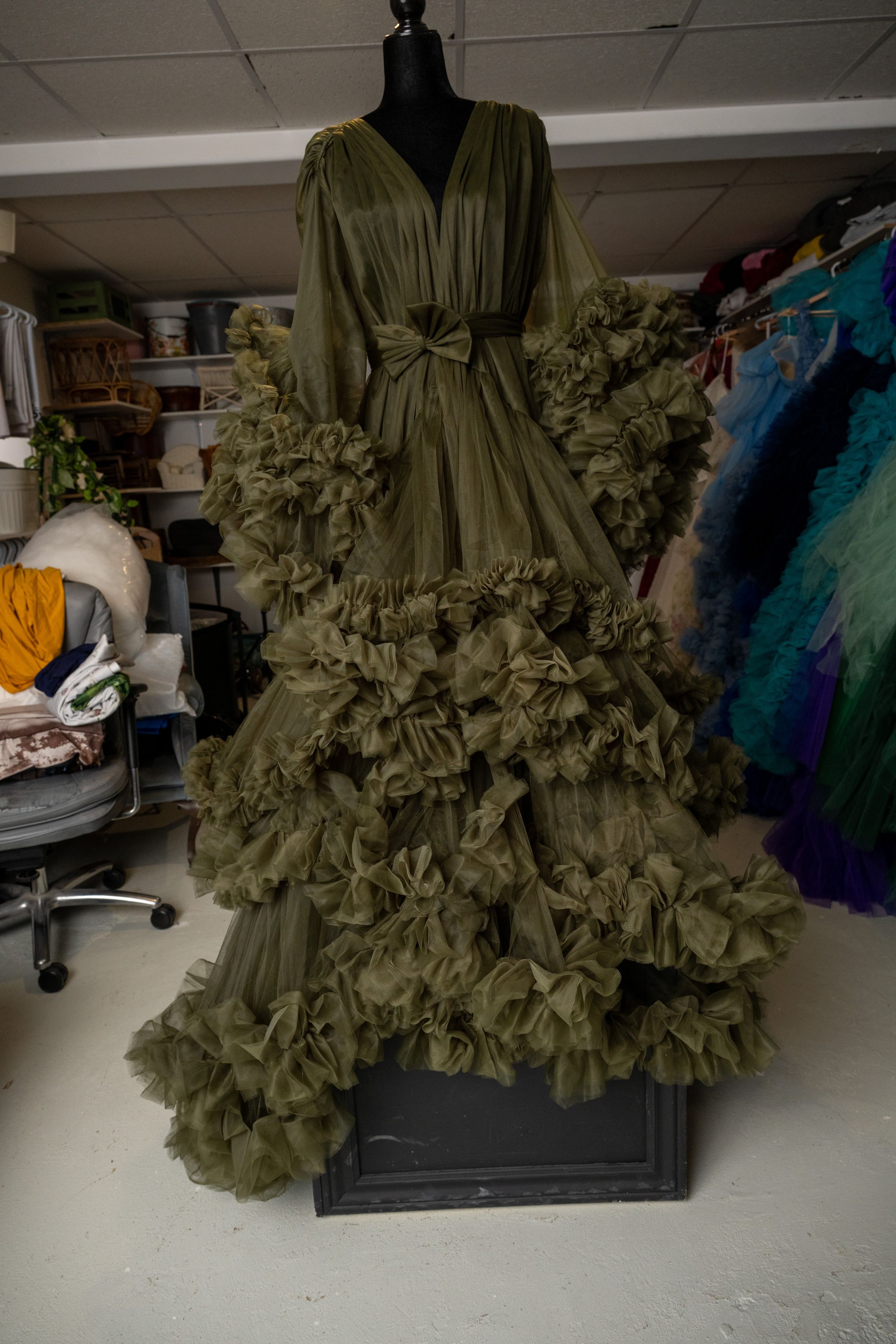 Dramatic olive green ruffled evening gown with voluminous tiered skirt and bow detail displayed on mannequin in studio.