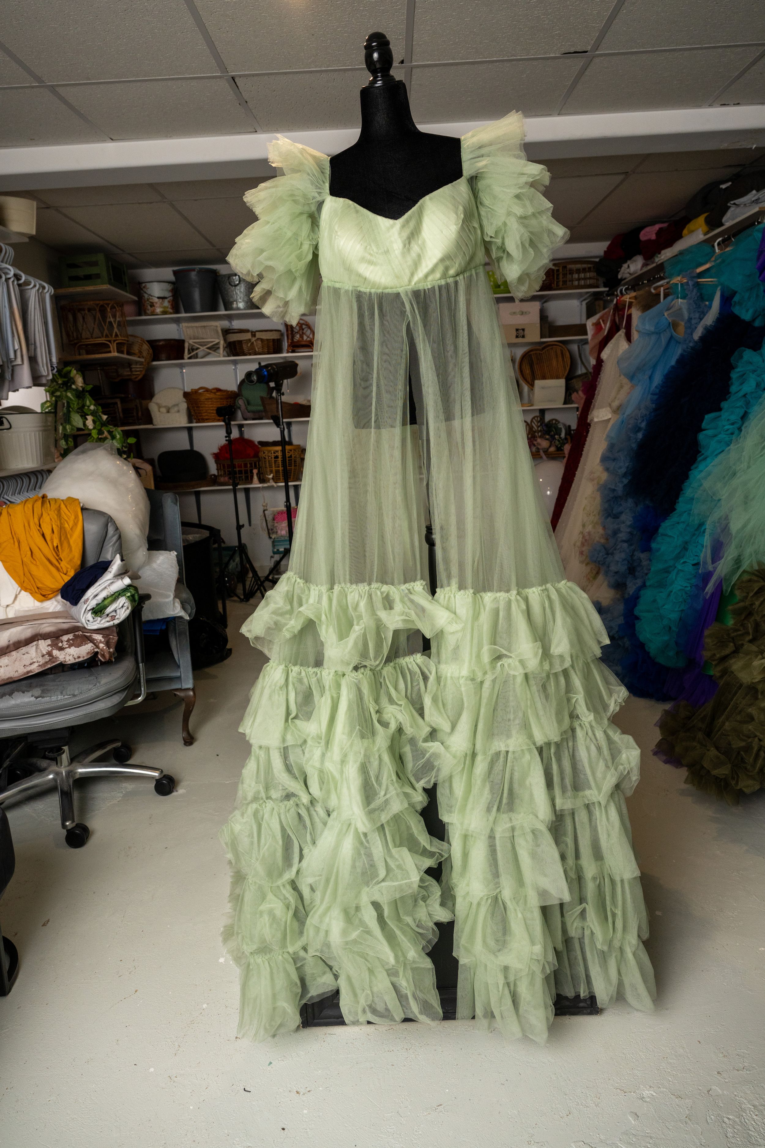 Ethereal mint green tulle gown with ruffled tiers and sheer overlay displayed on black dress form in workroom.