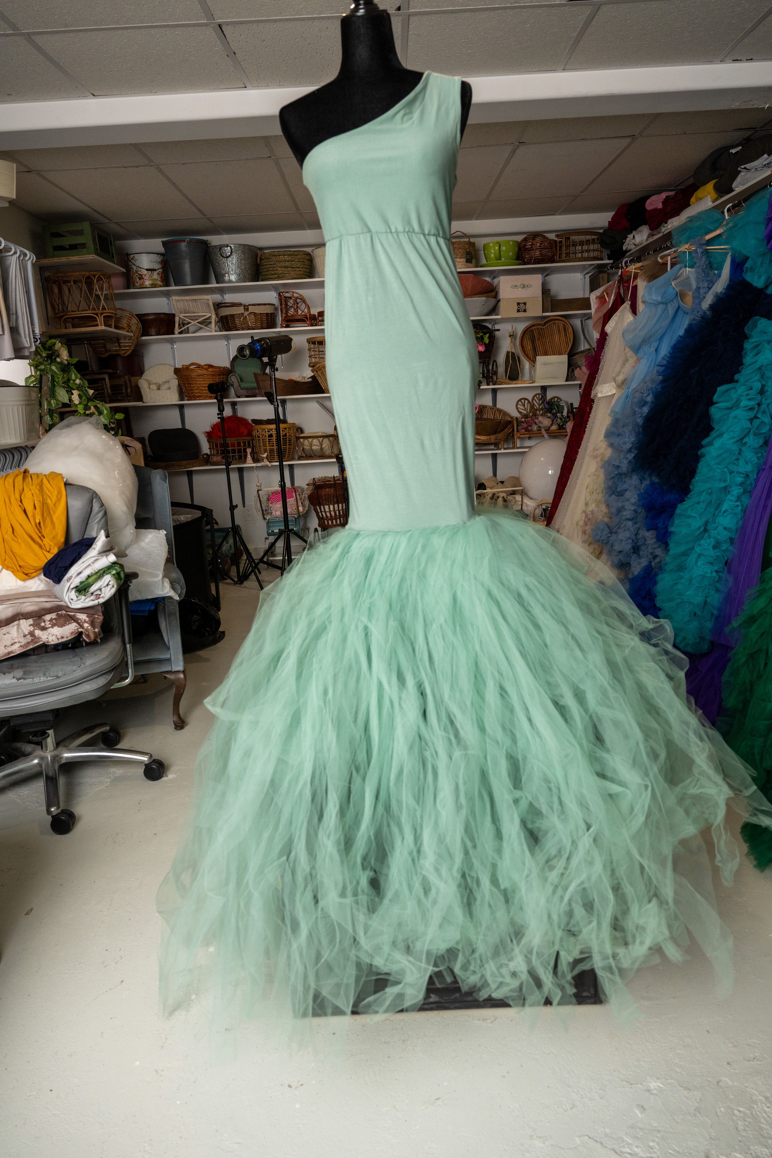 Mint green tulle ballgown with layered ruffles displayed in a studio setting with other formal dresses visible.
