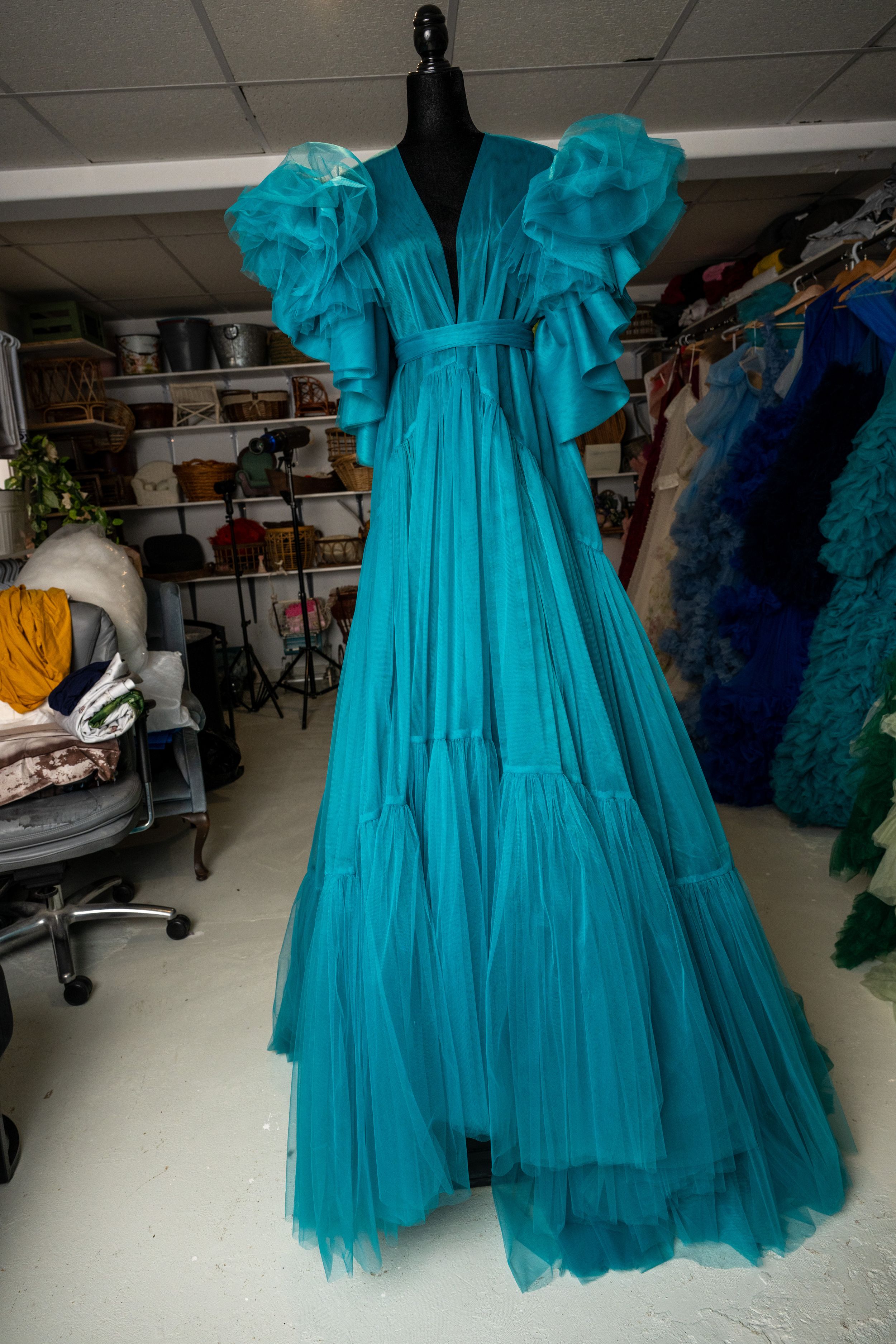 Turquoise tulle dress with ruffled layers displayed on a black mannequin in a storage room setting.