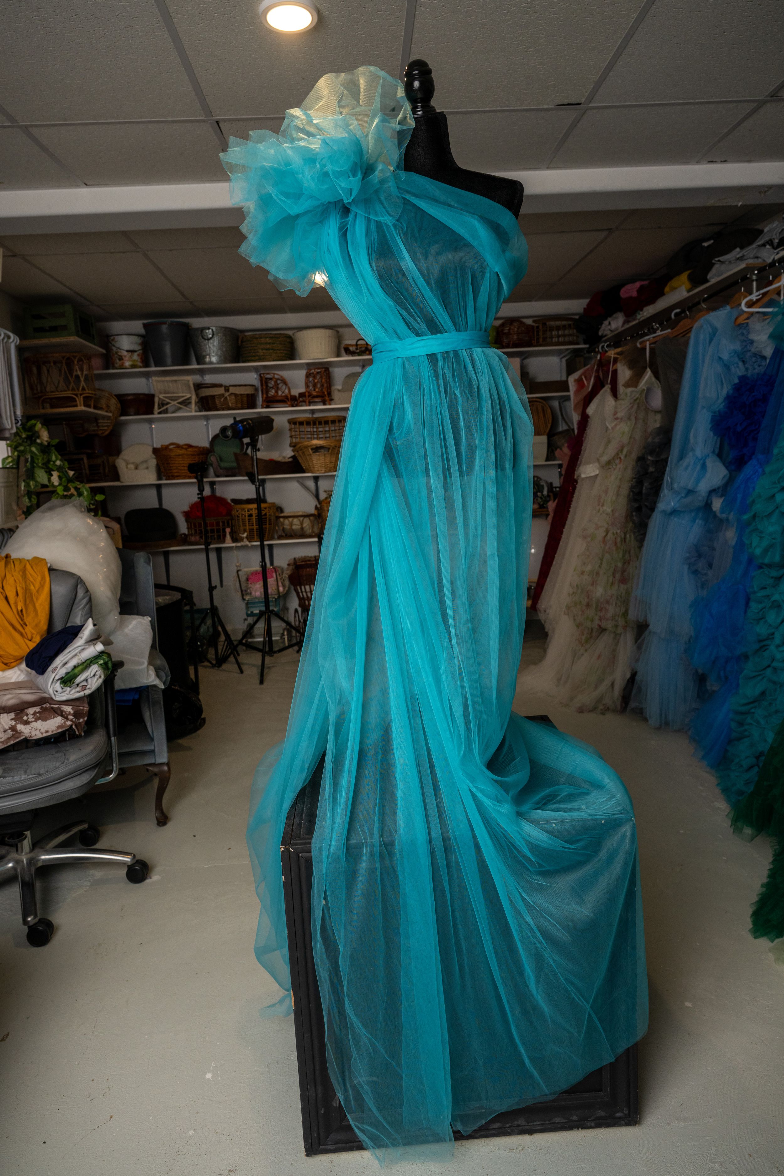 A turquoise tulle evening gown on display in a dress shop with other formal dresses visible in the background.