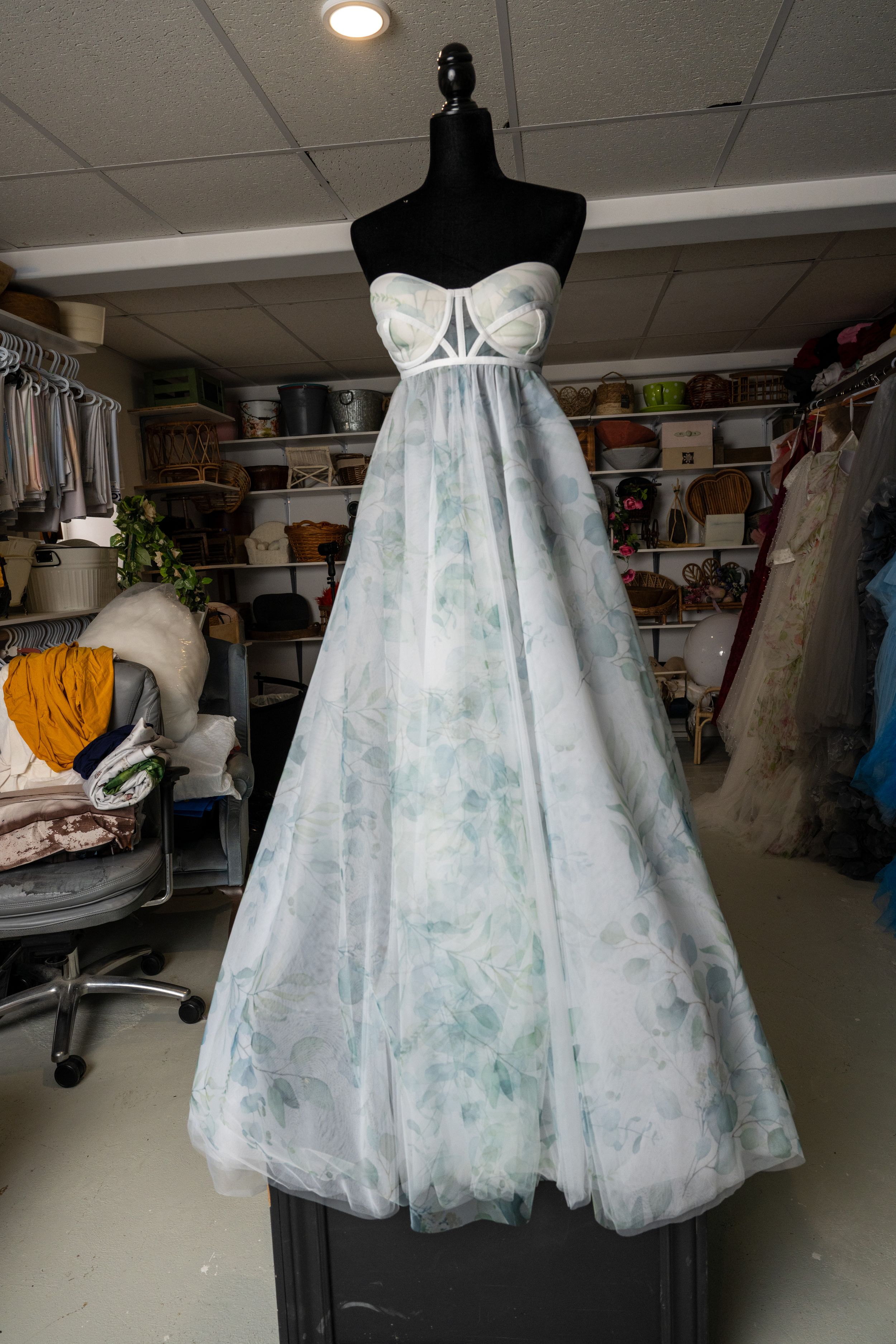 A white tulle wedding dress with light blue accents displayed on a mannequin in a studio workspace.