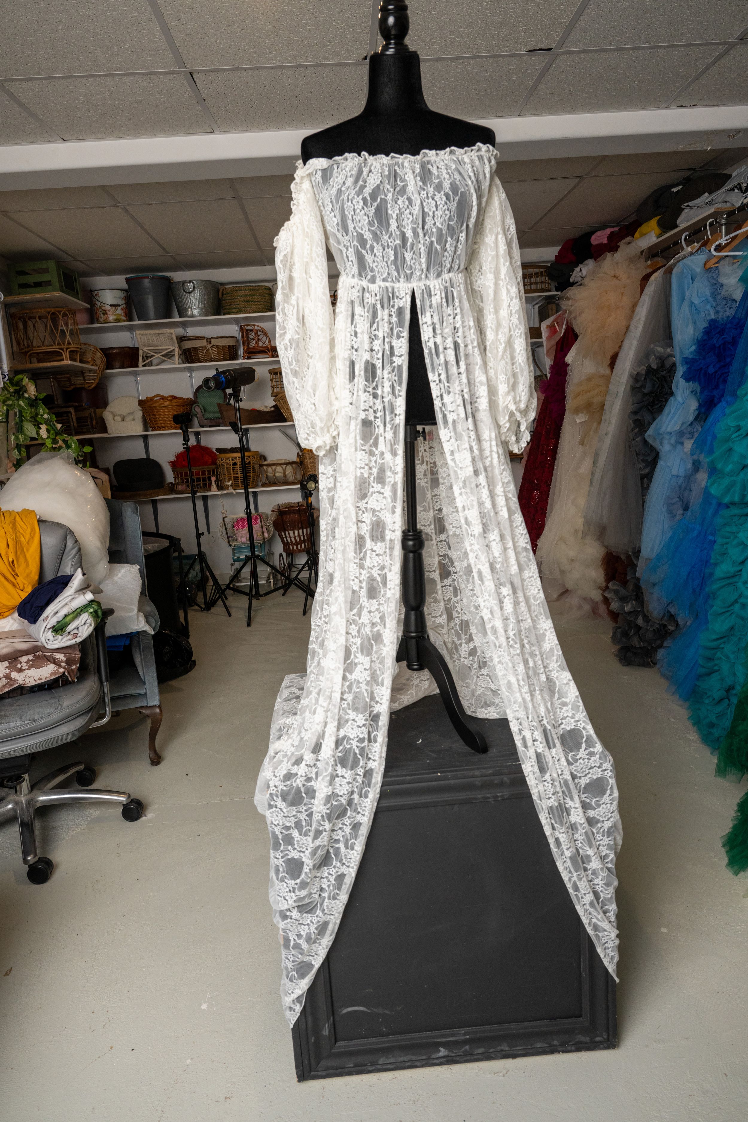 White lace fabric hangs from a clothing rack in a cluttered costume storage room with colorful garments visible.