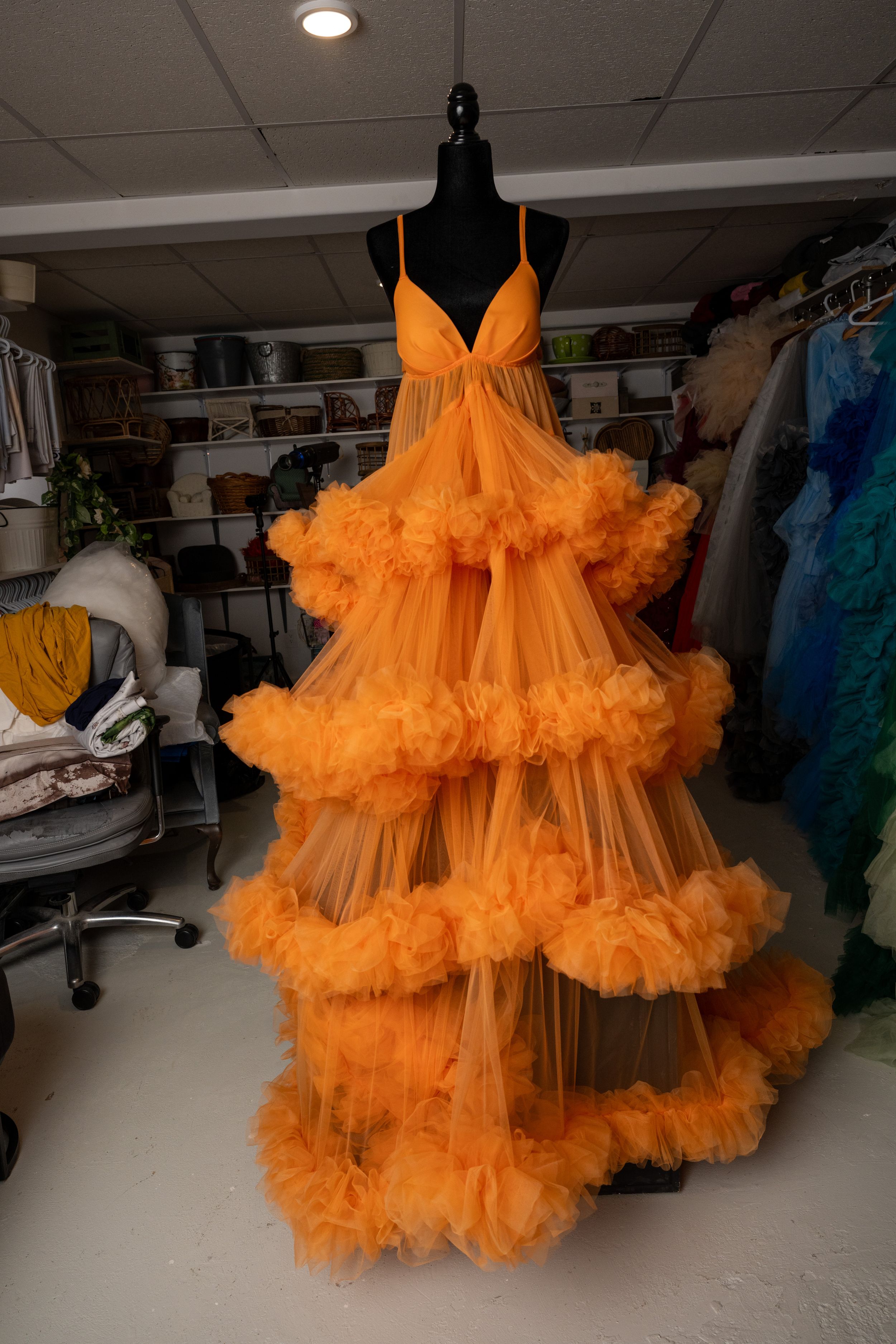 A vibrant orange tulle gown with dramatic ruffles and cloud-like layers displayed on a black mannequin in a studio setting.