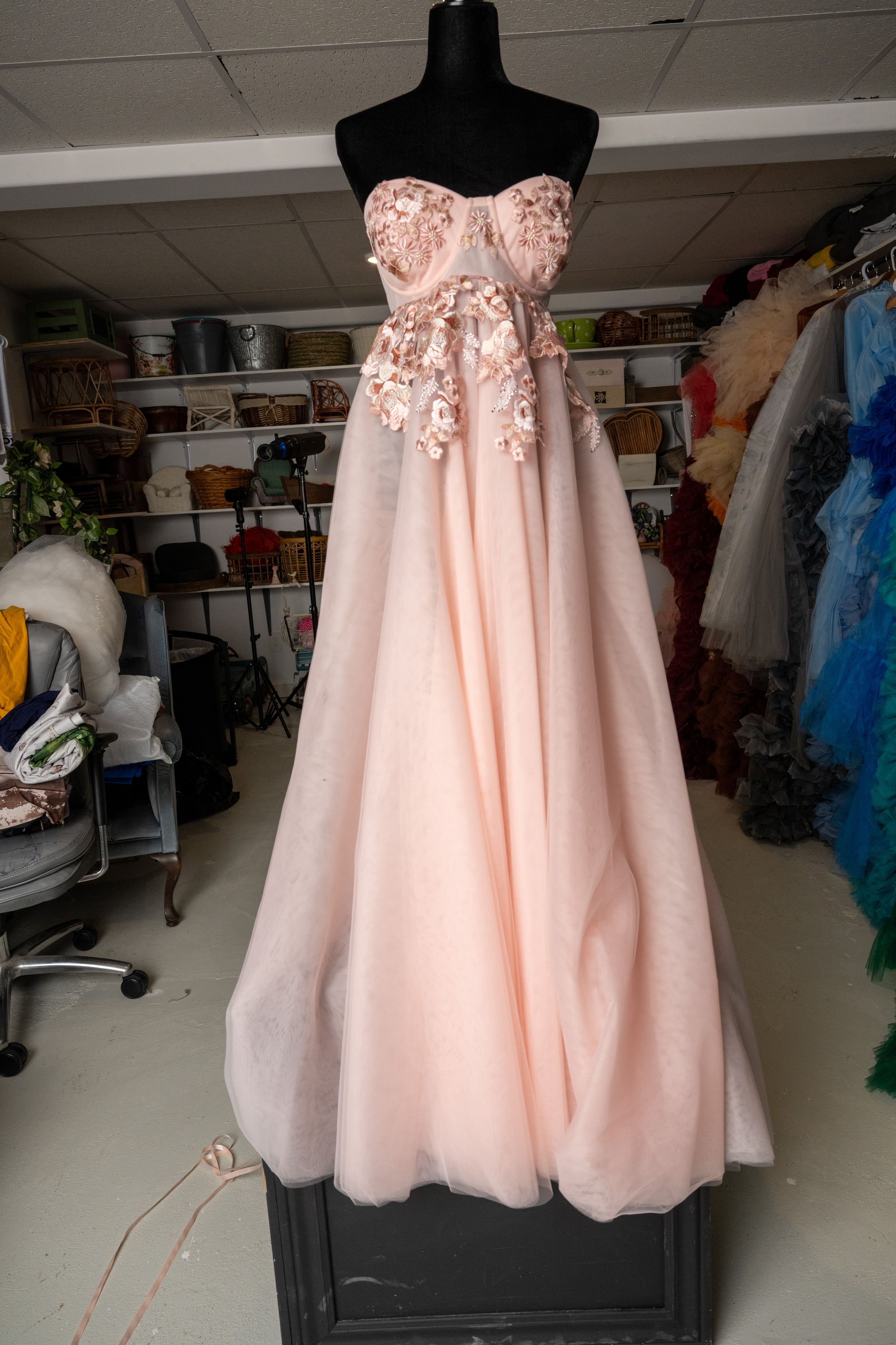 A blush pink strapless formal gown with floral embellishments displayed on a black mannequin in a dress shop.