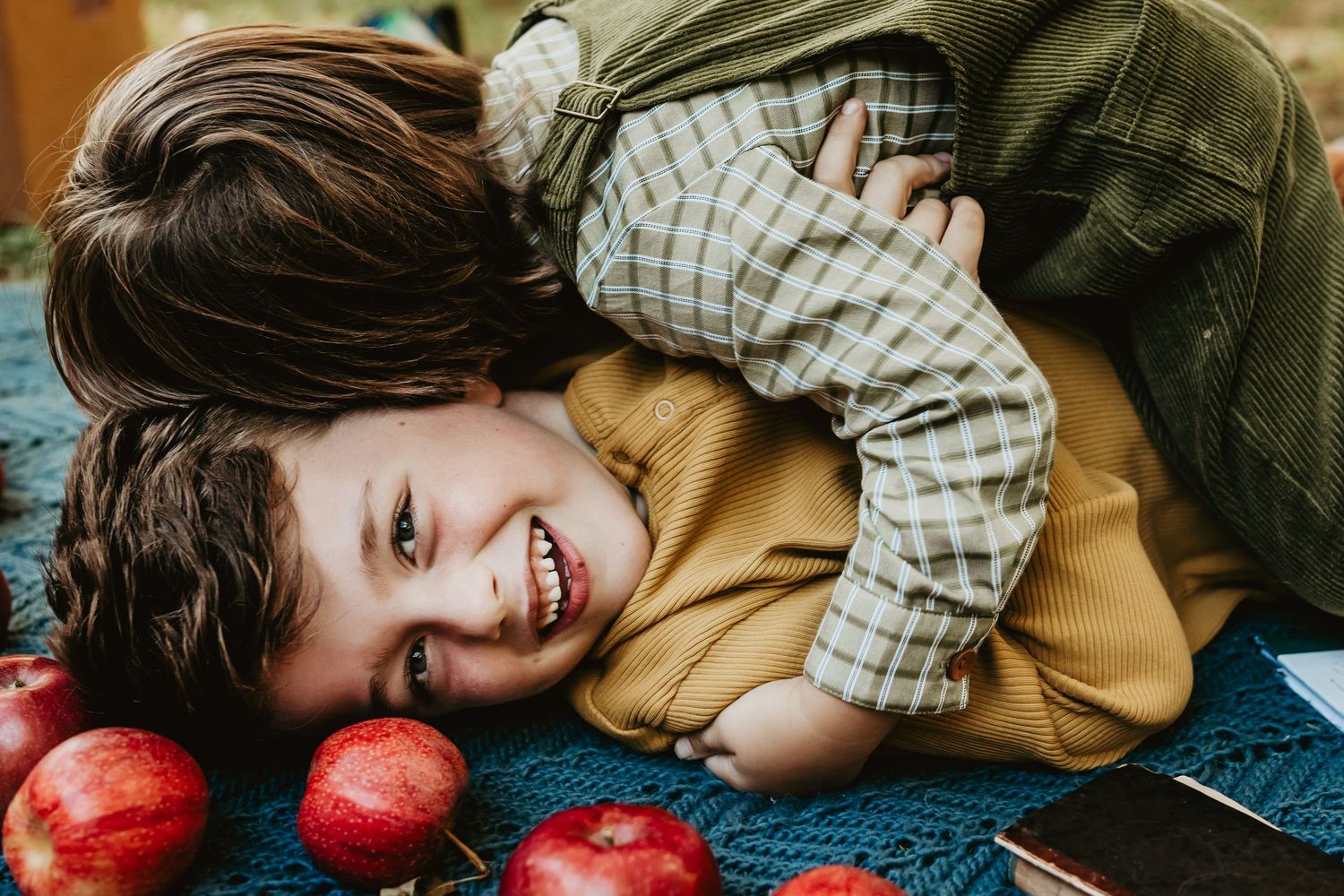 A child lies on a blue blanket next to bright red apples while smiling playfully.