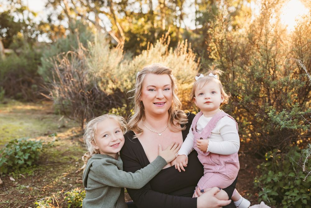 A joyful outdoor family portrait at golden hour with sunlight filtering through trees and foliage in the background.
