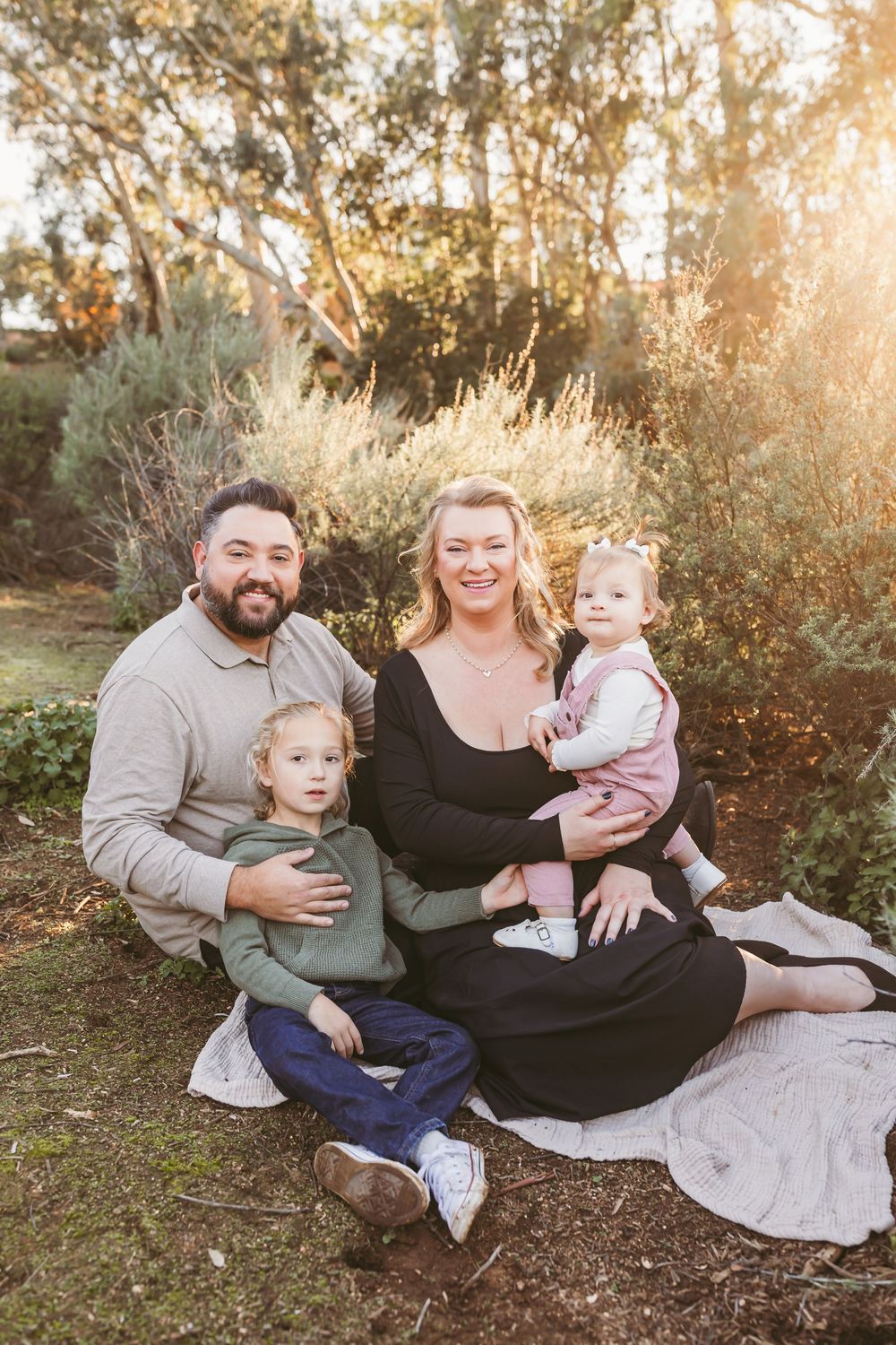 Family sitting together on a blanket outdoors during golden hour with trees and sunlight in the background.