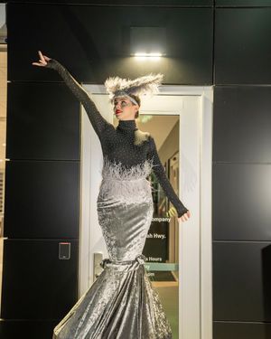 A performer in a glittering silver mermaid gown and feathered headpiece strikes a dramatic pose against a dark wall.