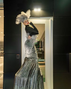 Performer in a glamorous silver gown and feathered headpiece poses dramatically in a doorway.