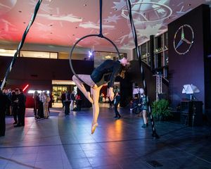 An aerial acrobat performs on a hoop in front of a Mercedes-Benz logo at an elegant event space.