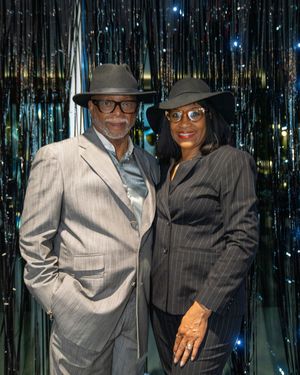Two people in formal attire with hats pose together against a sparkly backdrop at an event.