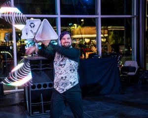 A performer in a floral shirt dances energetically at night in front of illuminated neon light decorations.