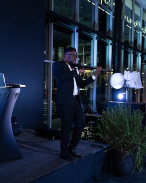 Performer in formal attire sings on stage with lighting equipment and glass windows in background.
