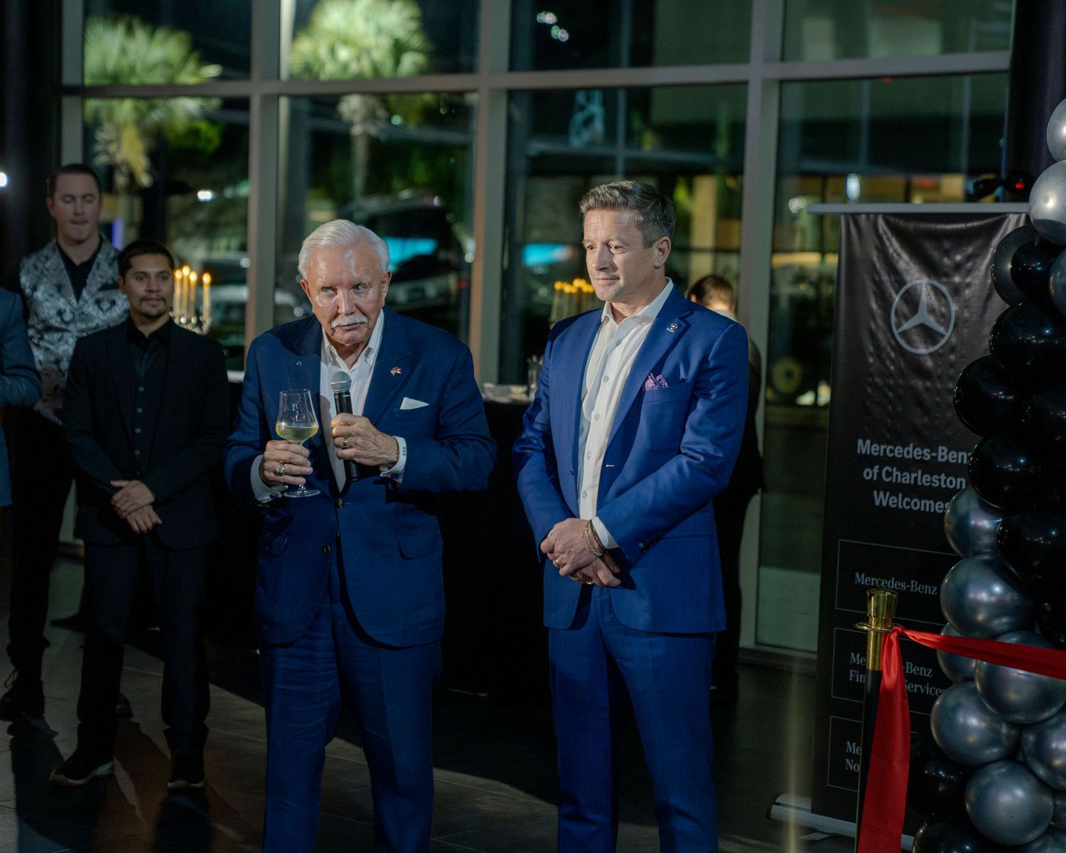 People in blue suits stand together at an event with glass windows and red ropes visible in the background.