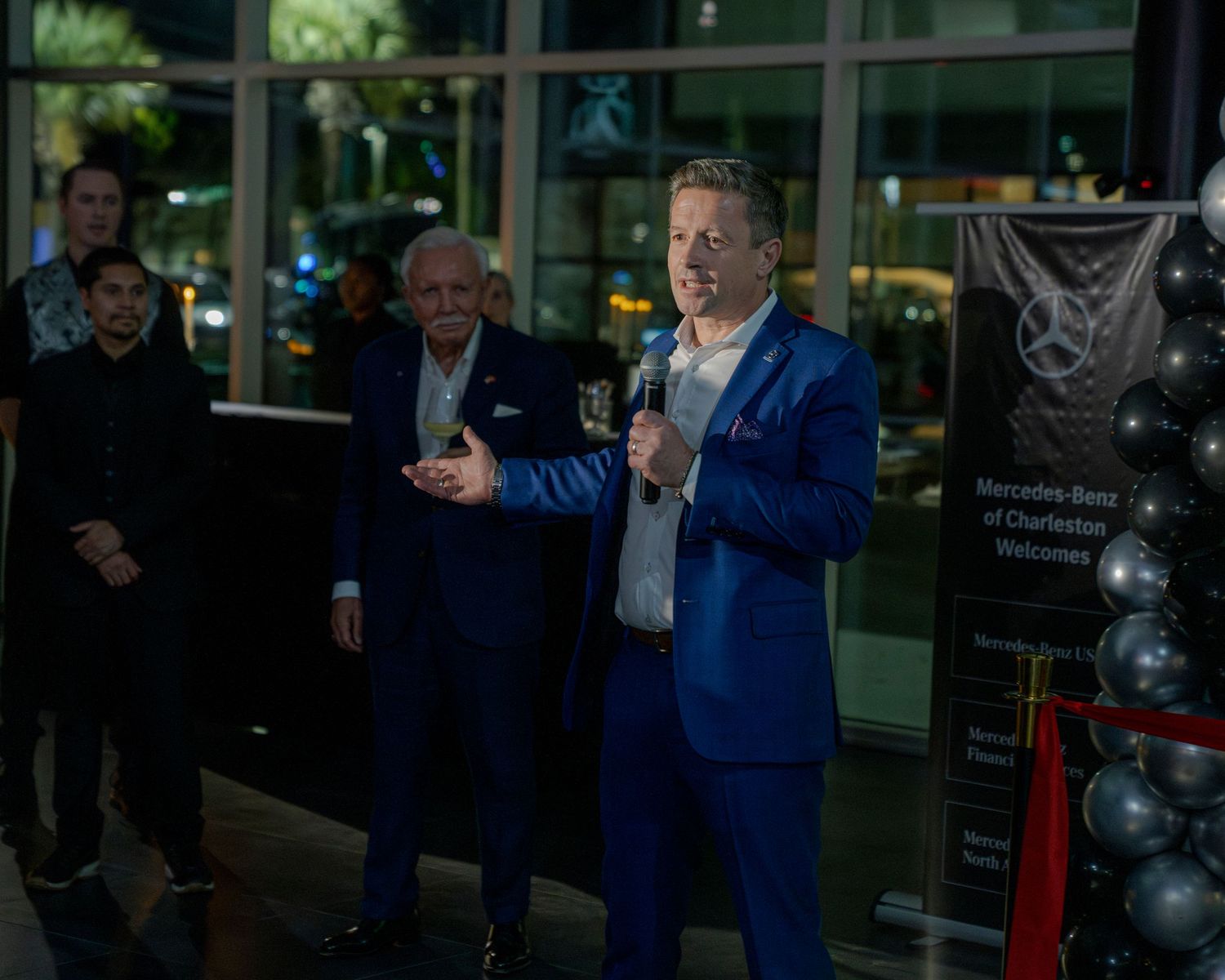 People in blue suits stand together at an event with glass windows and red ropes visible in the background.