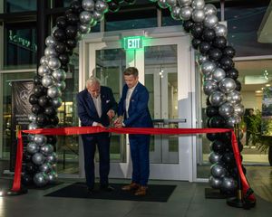 A ribbon cutting ceremony takes place under a decorative silver balloon arch at a building entrance.