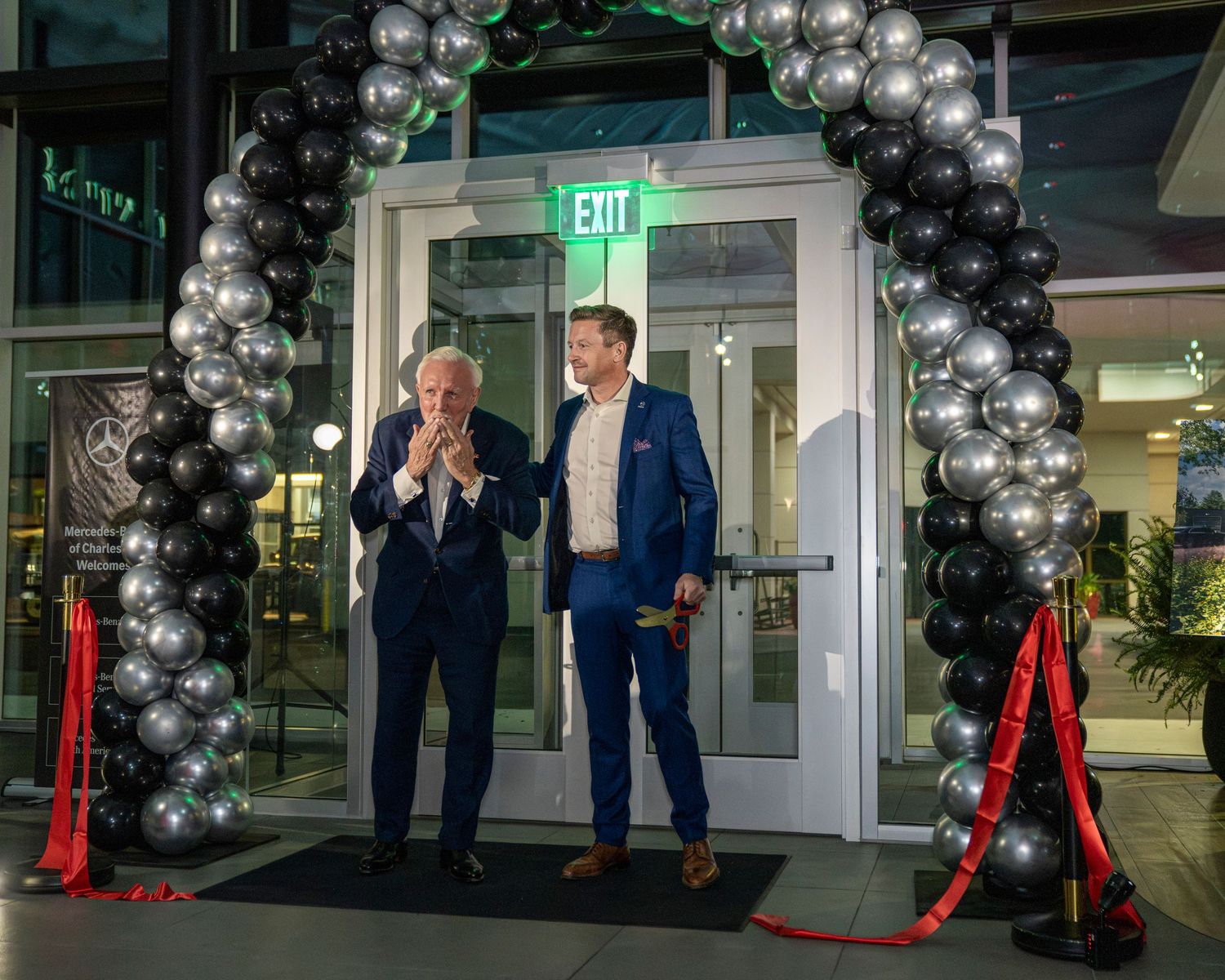 Two people stand under a silver and black balloon arch at a nighttime event entrance with an exit sign visible.