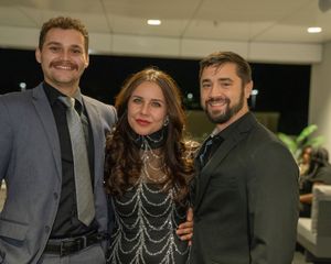 Three guests in formal attire smile together at an evening event.