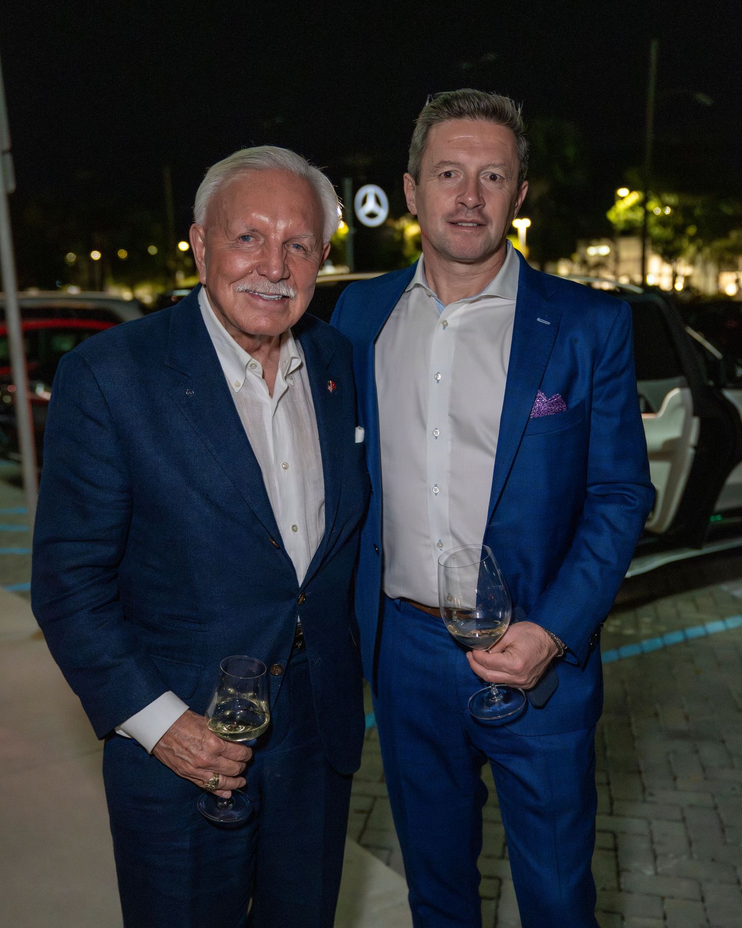 Two men in matching blue suits smile while holding wine glasses at an evening event.