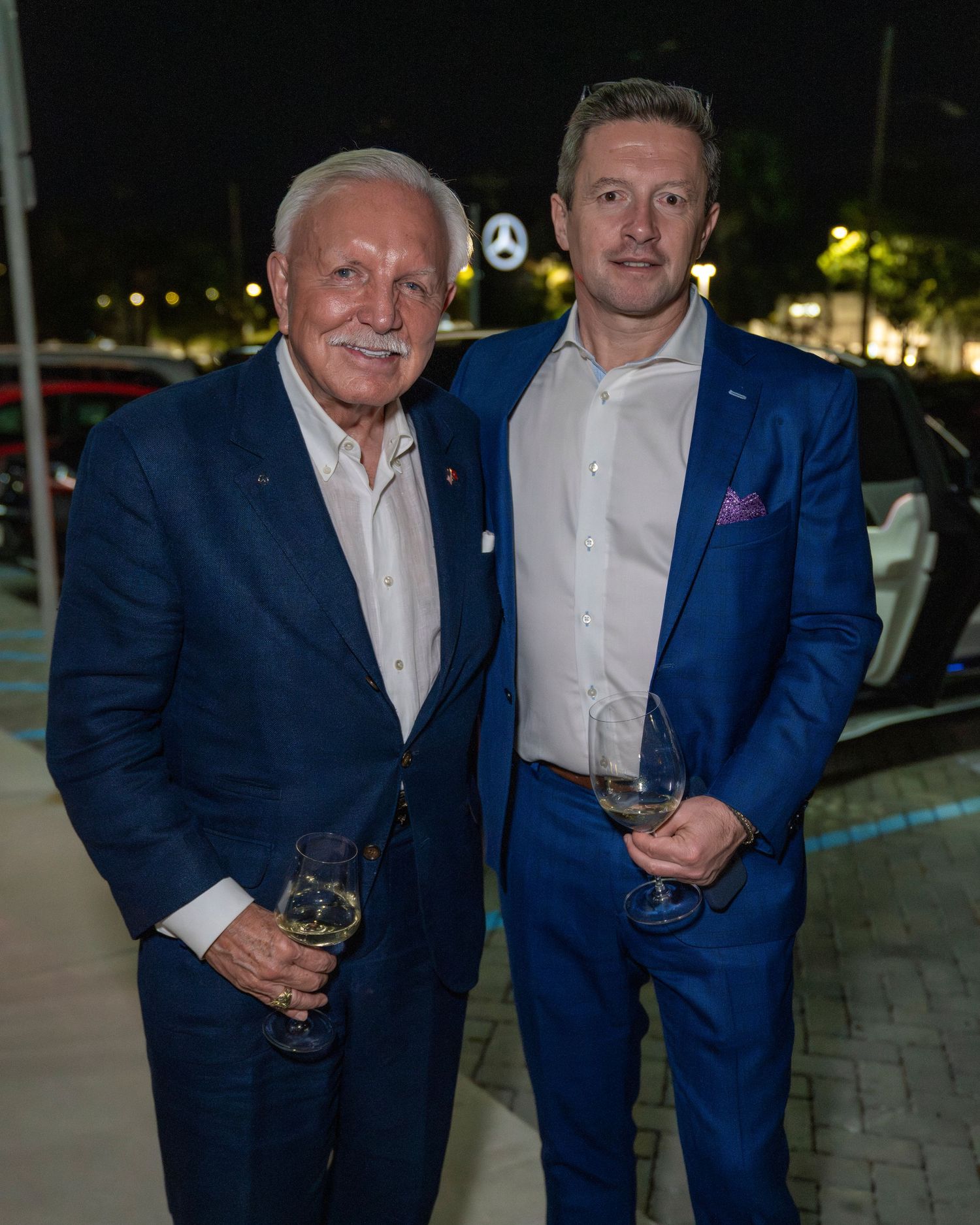 Two men in navy blue suits and white shirts pose together at an evening event with wine glasses.