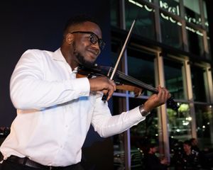 A violinist performs on stage wearing a white shirt against a dark background.