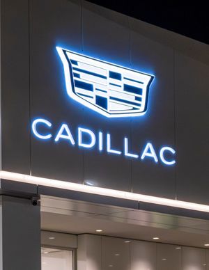 Illuminated blue Cadillac logo and text signage mounted on dark wall of car dealership at night.