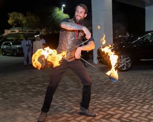 Fire performer spinning flaming batons in dramatic nighttime display outside building.