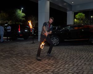 Fire performer spinning flaming batons in a dark parking area with cars in the background.