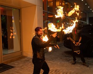 Fire performer spins flaming batons outside a building entrance at night.