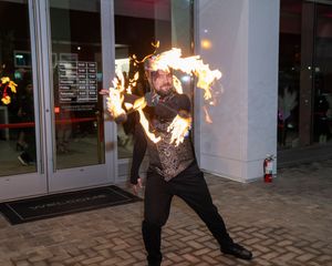 Fire dancer performing with flaming props against a modern building facade creating dramatic light effects.